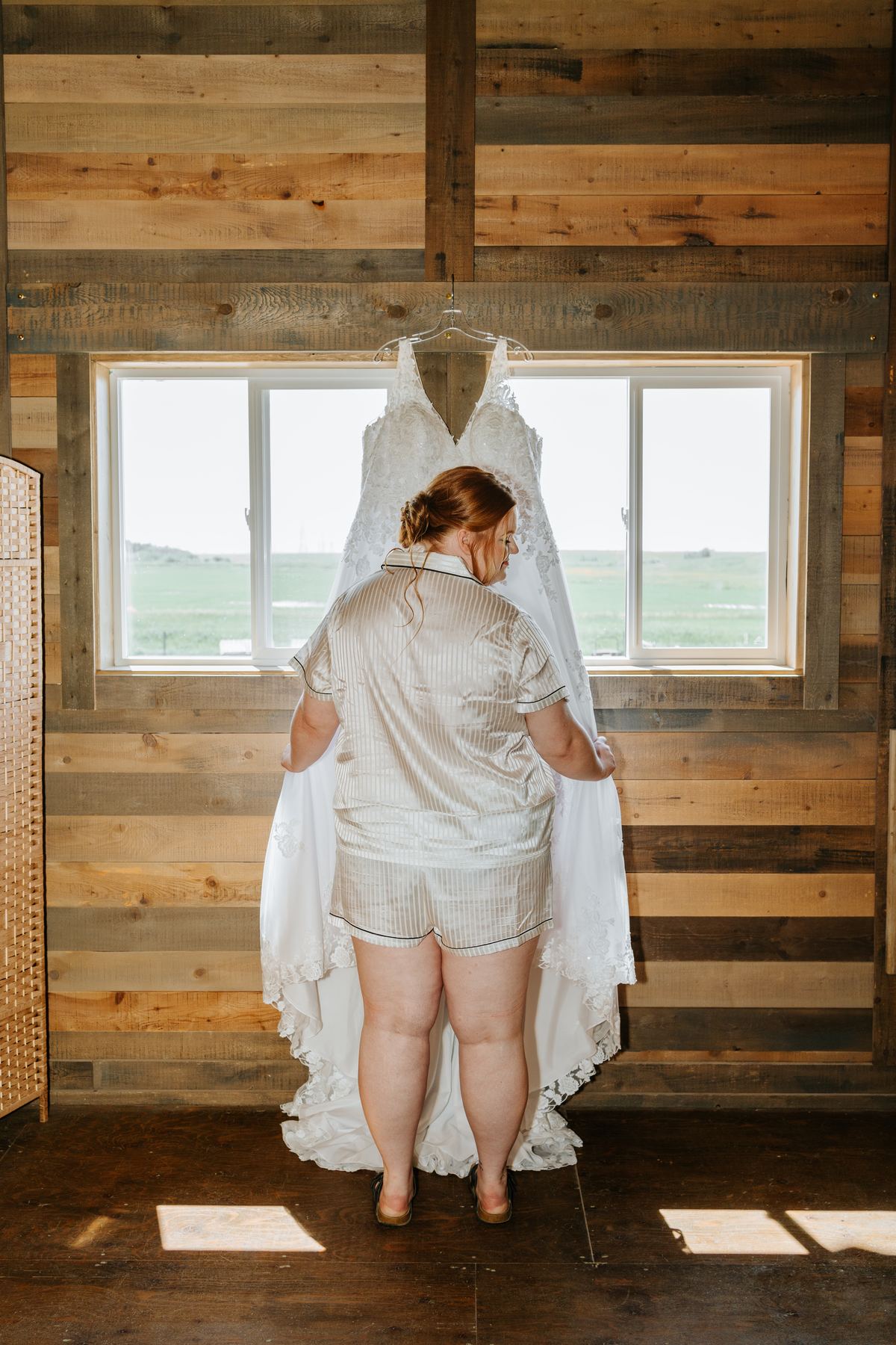 Bride Alysia in satin pyjamas looking at her wedding gown hanging in the barn window — Deer Meadow Farm Manitoba