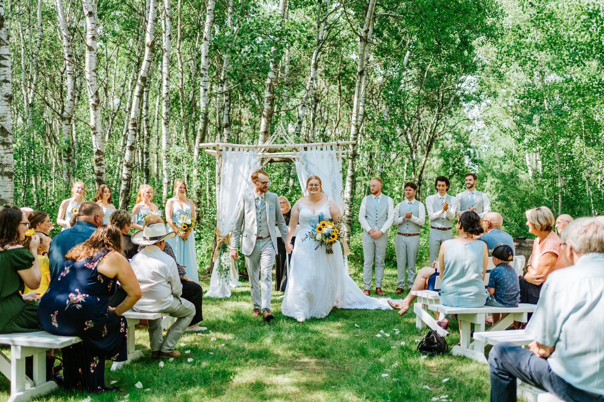 Wide ceremony view — Alysia and Josh at the log arbor with guests filling white benches in the birch forest at Deer Meadow Farm Manitoba