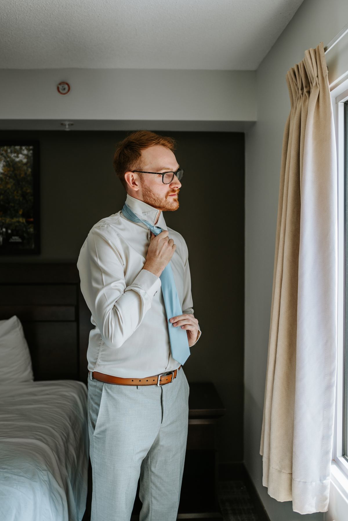 Groom Josh adjusting his blue tie in the hotel room window light — Deer Meadow Farm wedding Manitoba by Ngo Photography