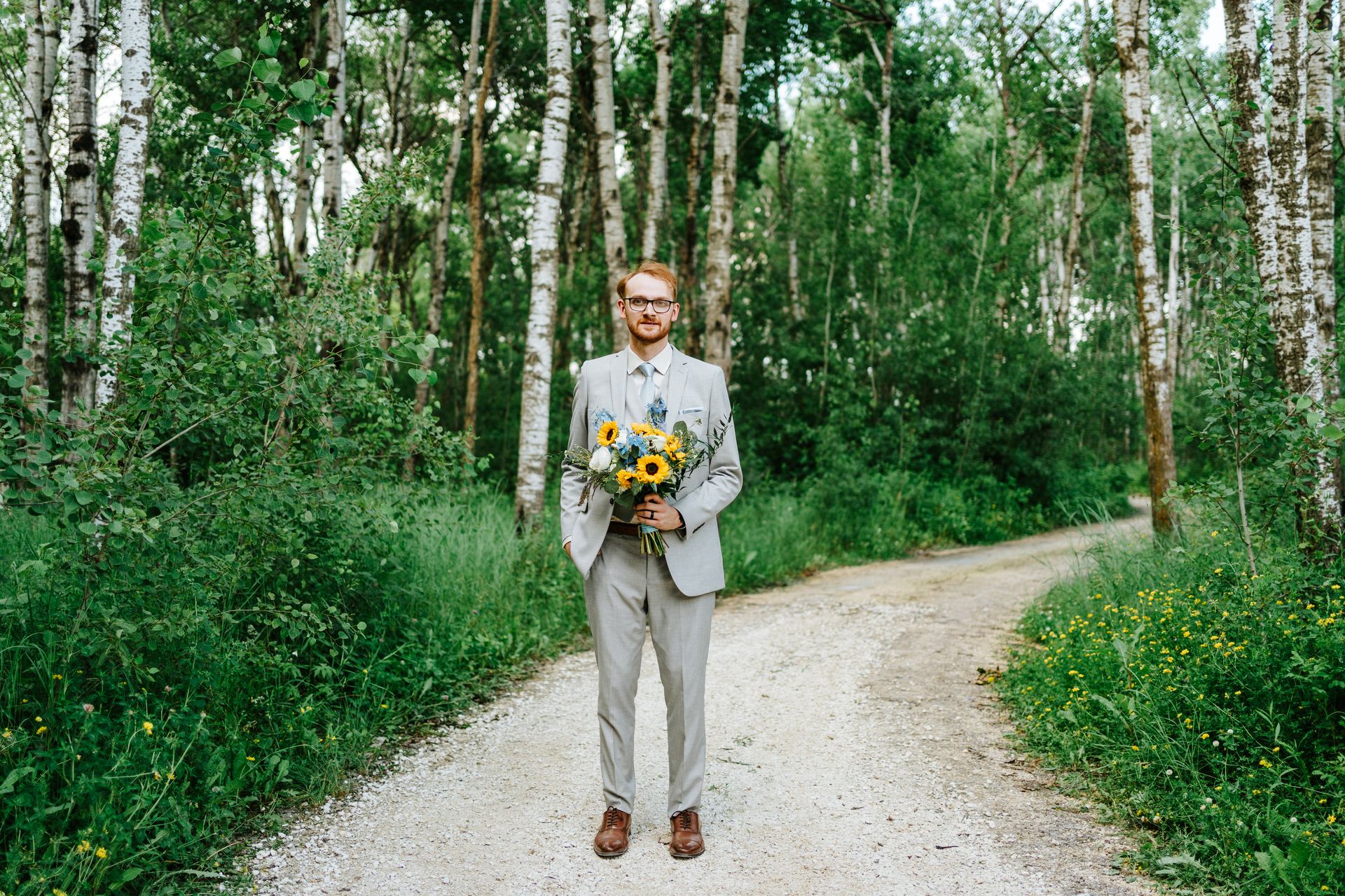 Groom Josh standing alone on the birch forest path with sunflower bouquet — Deer Meadow Farm Manitoba wedding