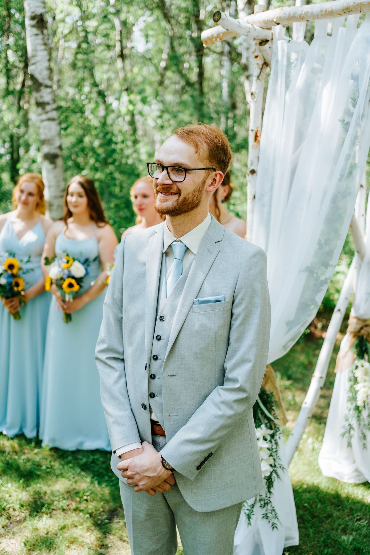 Groom Josh smiling at the log arbor as bridesmaids arrive — Deer Meadow Farm birch forest ceremony Manitoba