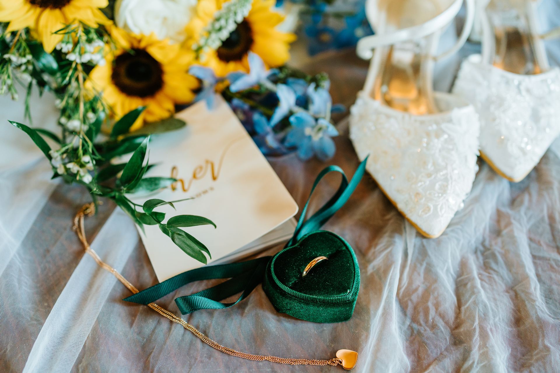 Bridal flat lay at Deer Meadow Farm — green velvet ring box, vow book, sunflower bouquet and lace shoes on sheer fabric