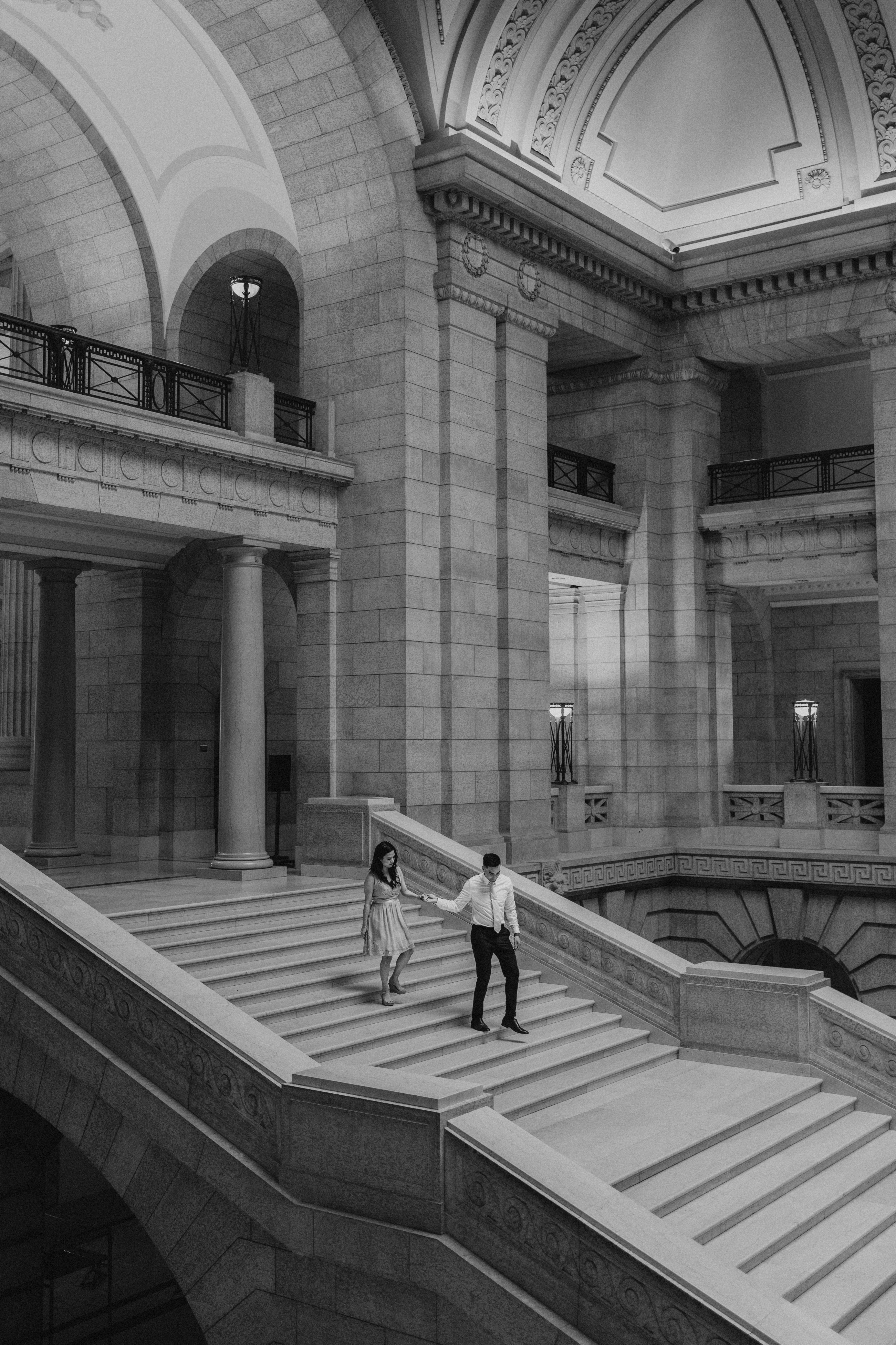 Black and white architectural photo of AJ and Julie walking on the grand staircase inside the rotunda of the Manitoba Legislative Building Winnipeg — Ngo Photography