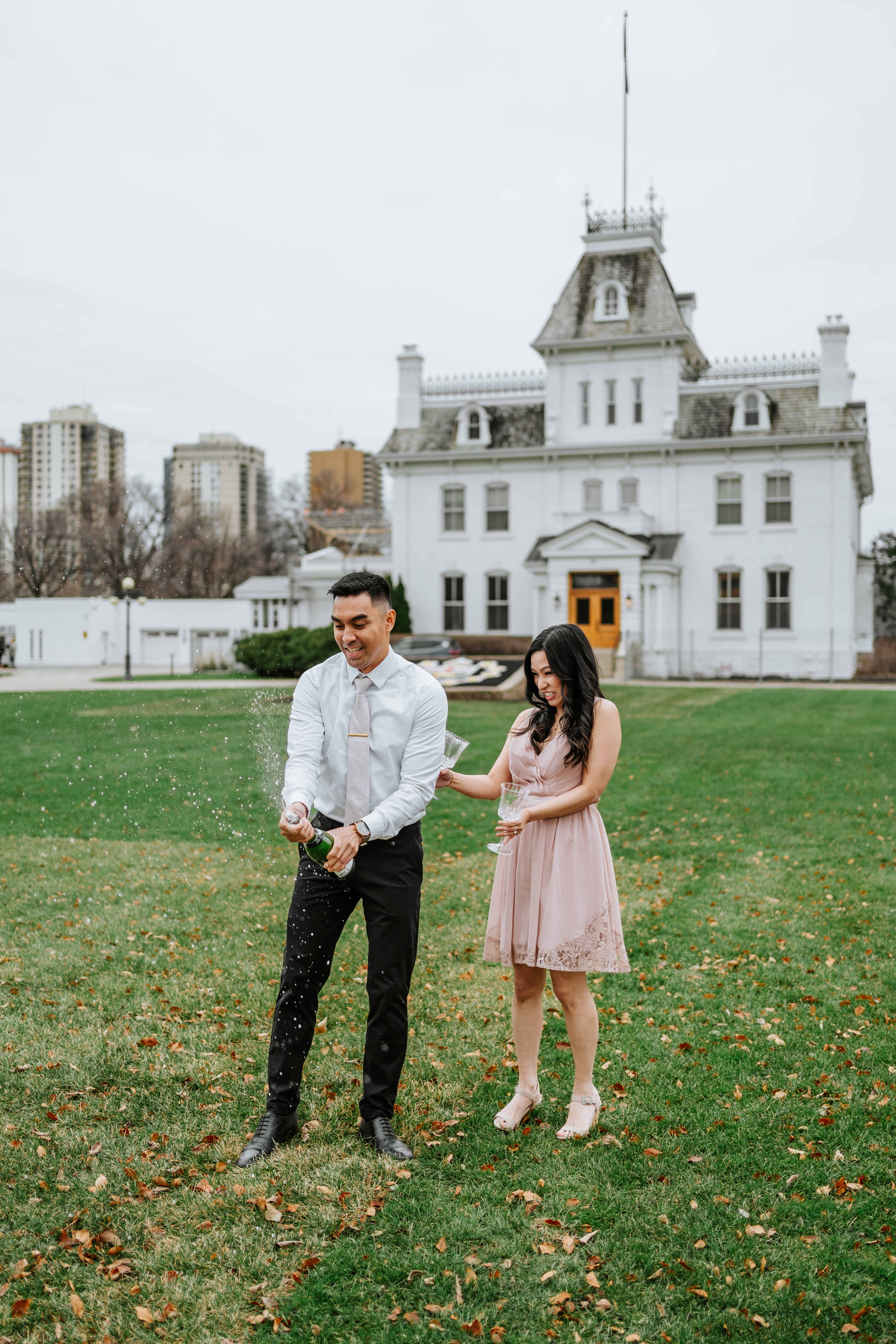 AJ and Julie popping champagne on the south lawn of the Manitoba Legislative Building with Government House behind them Winnipeg — engagement photography by Ngo Photography