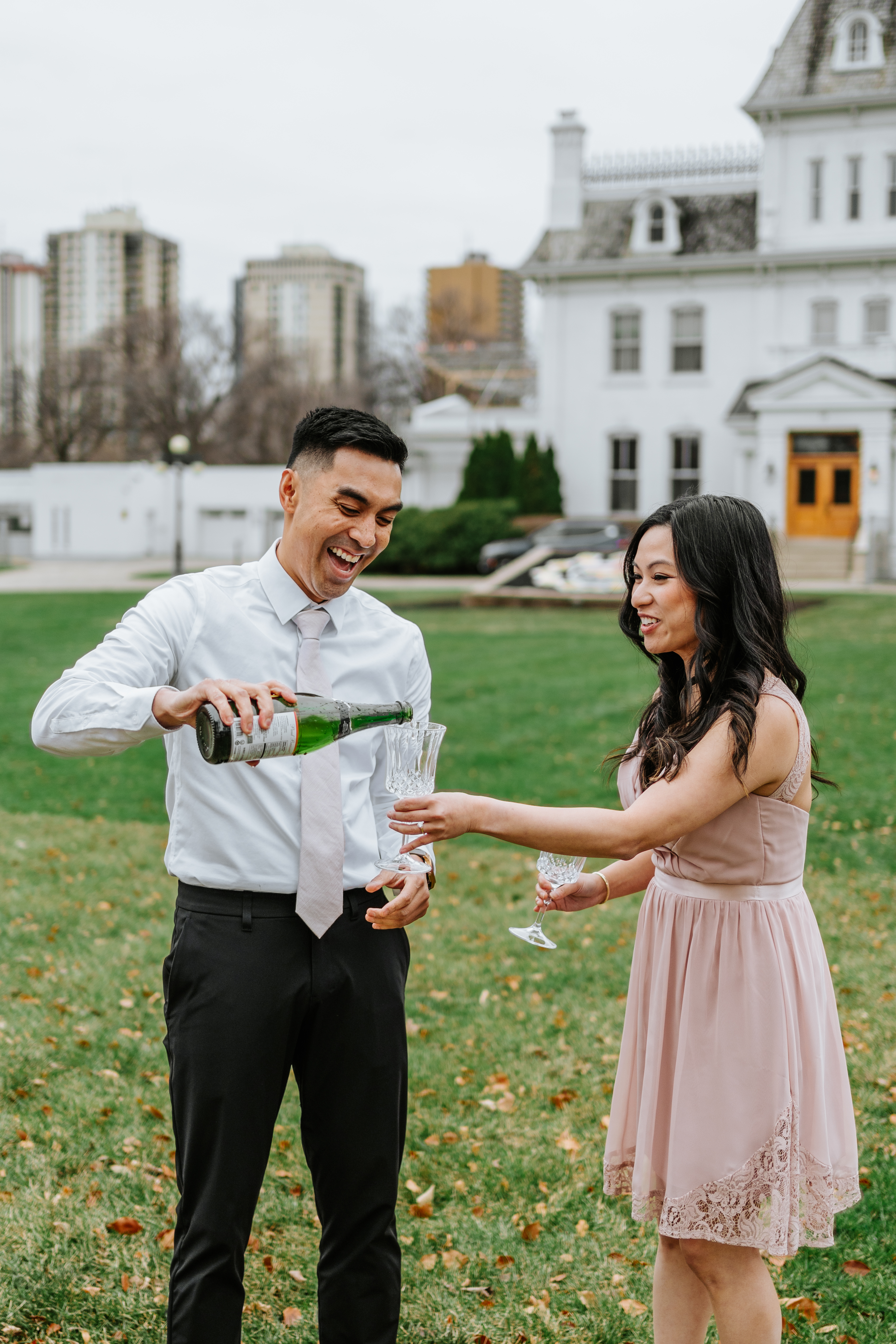 AJ laughing while pouring champagne for Julie on the Manitoba Legislative grounds with historic Government House in background — engagement session by Chris Ngo Winnipeg