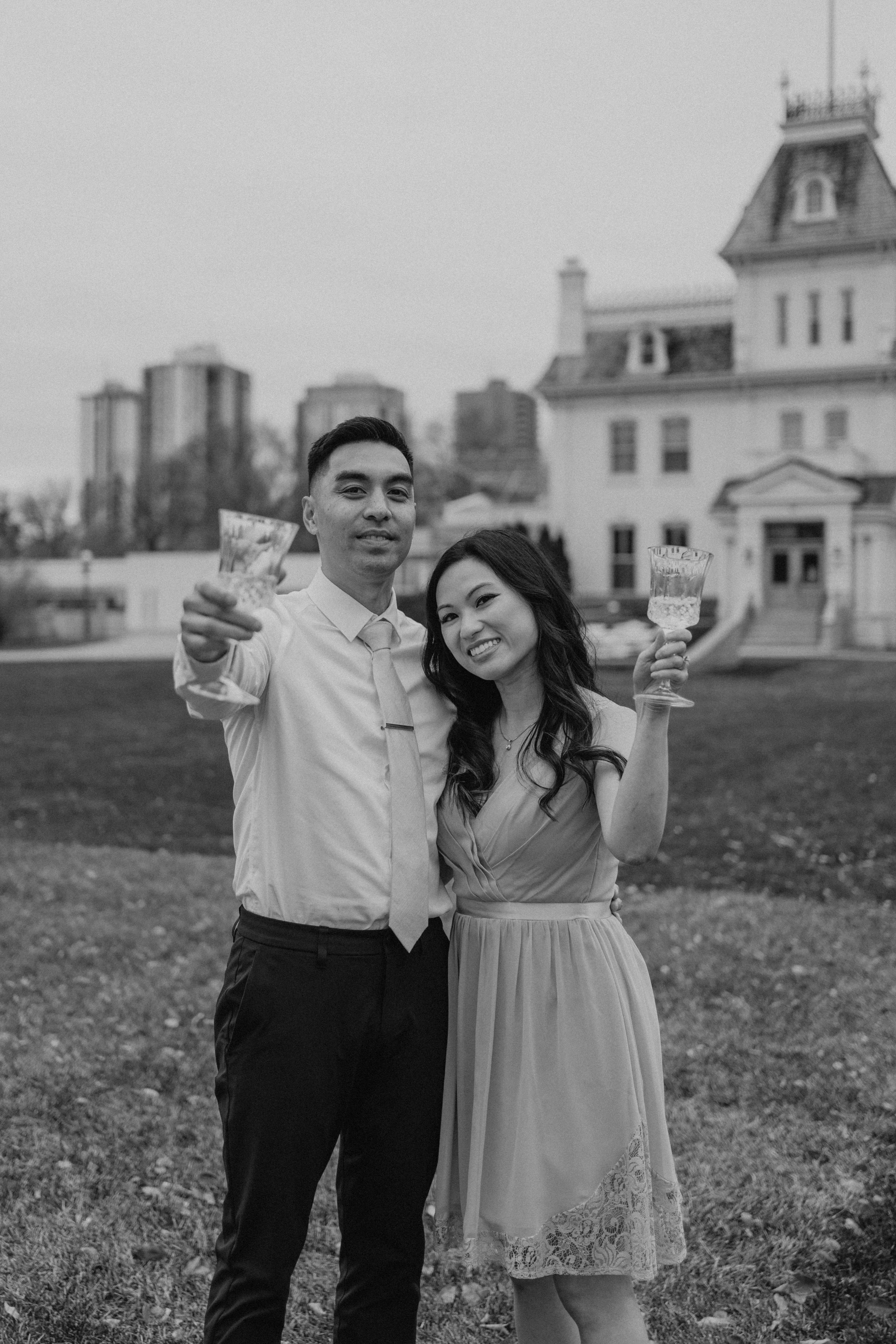 Black and white photo of AJ and Julie toasting champagne glasses on the Manitoba Legislative Building grounds Winnipeg — engagement photography by Ngo Photography