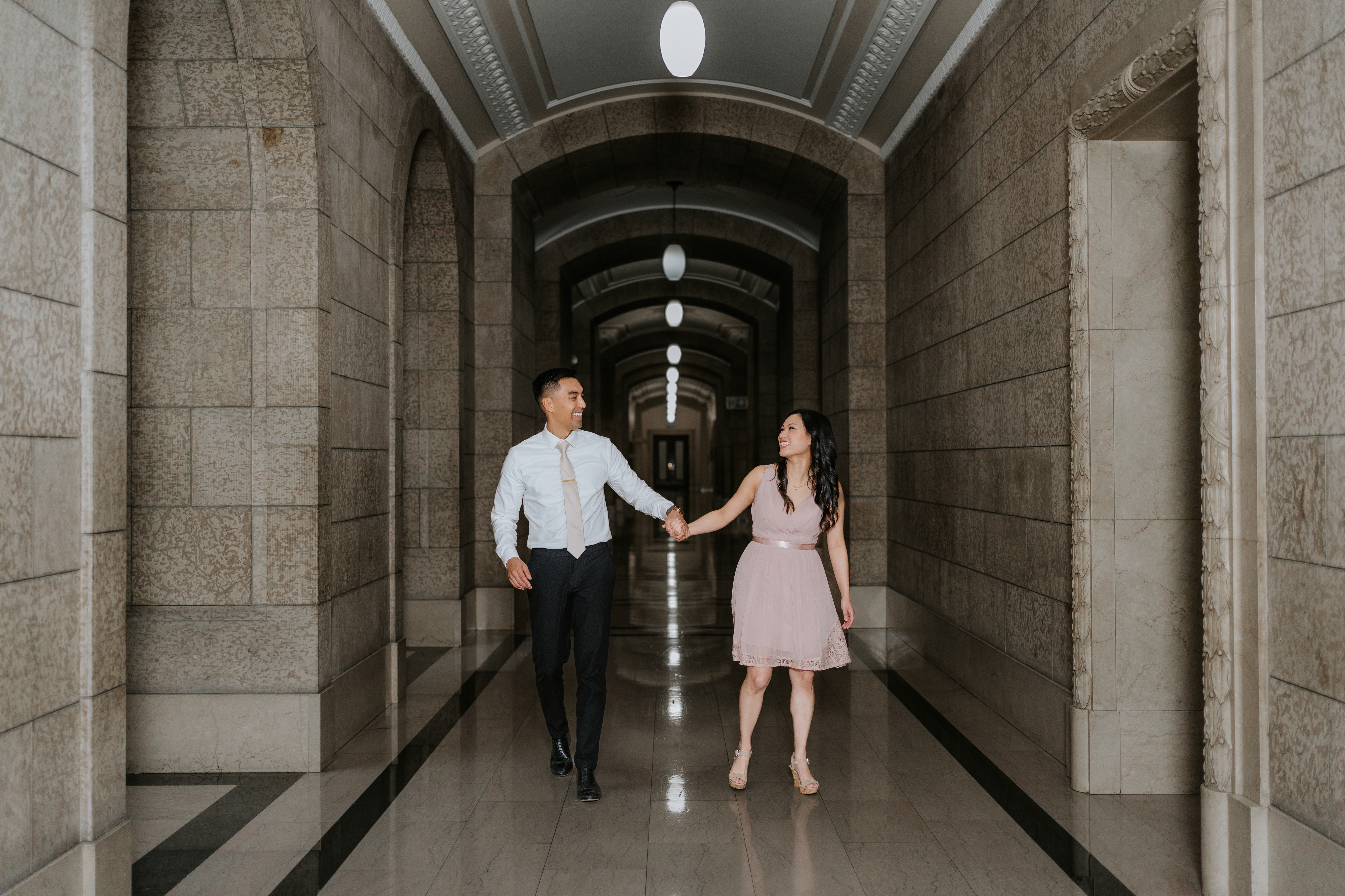 AJ and Julie walking hand in hand through a long arched corridor at the Manitoba Legislative Building Winnipeg — engagement photography by Chris Ngo Ngo Photography