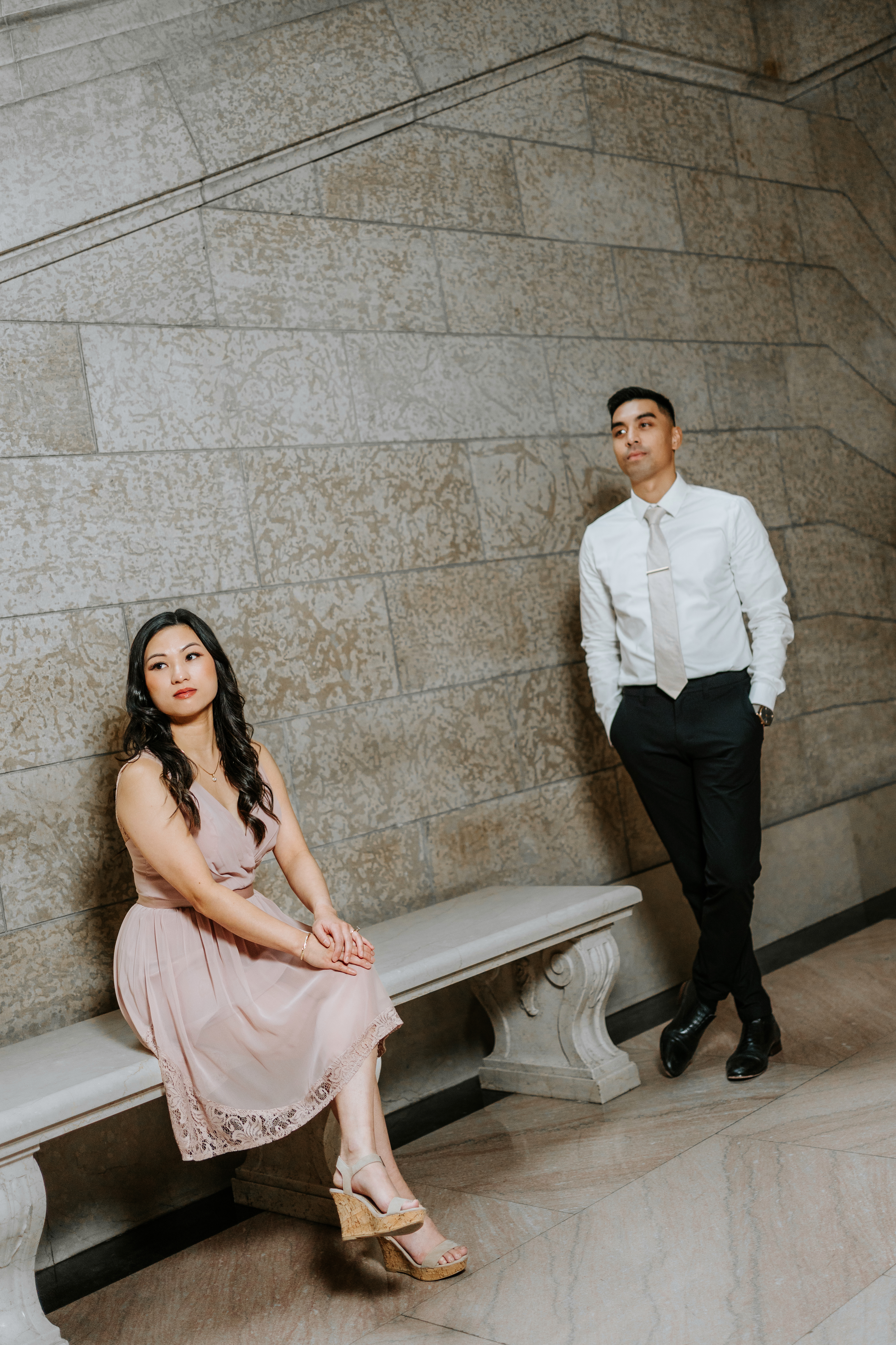 Editorial engagement portrait of Julie seated and AJ standing by stone bench at the Manitoba Legislative Building Winnipeg — engagement photography by Chris Ngo
