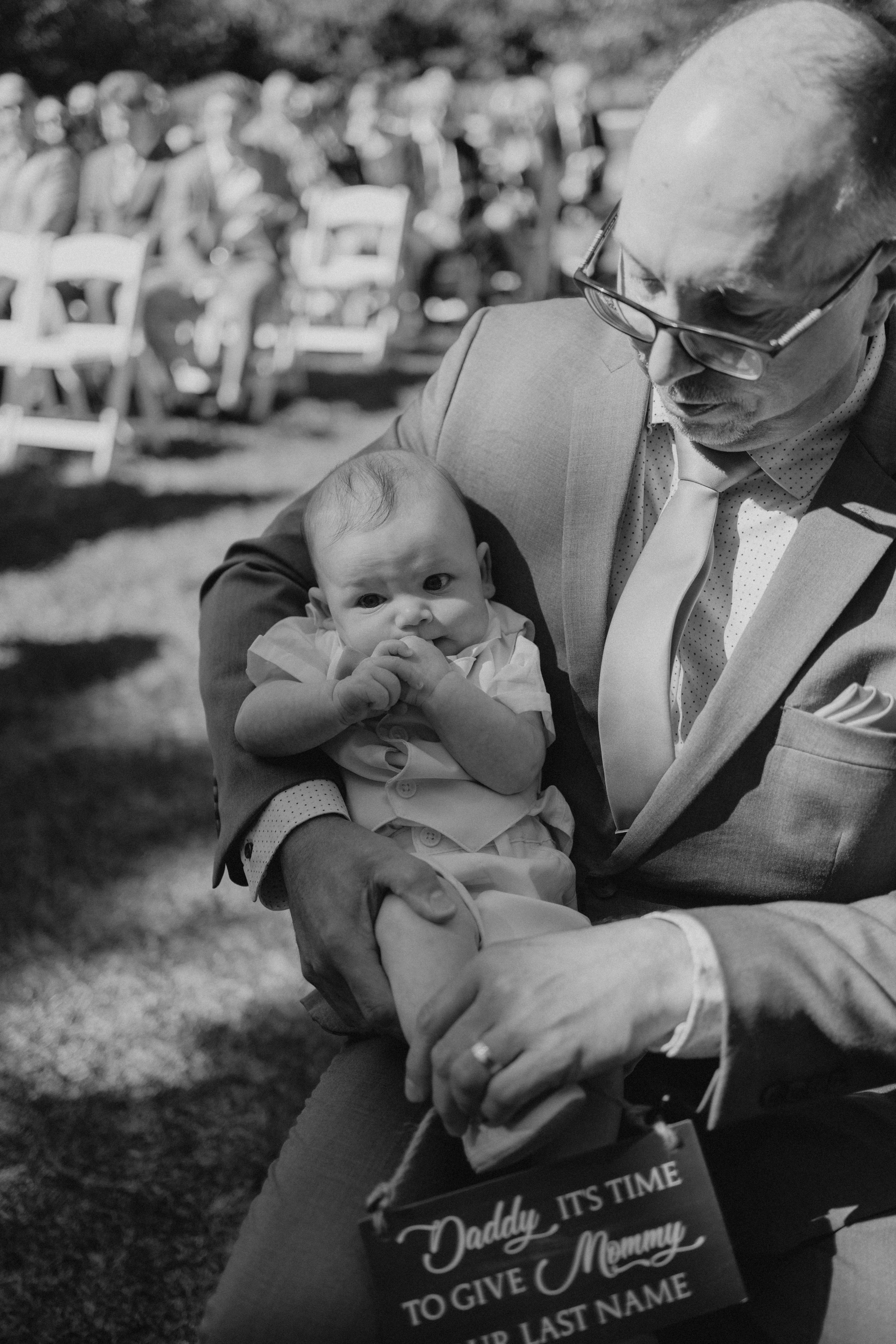 Baby held by grandfather at Ashgrove Acres ceremony with sign reading Daddy it's time to give Mommy our last name — wedding photography by Chris Ngo