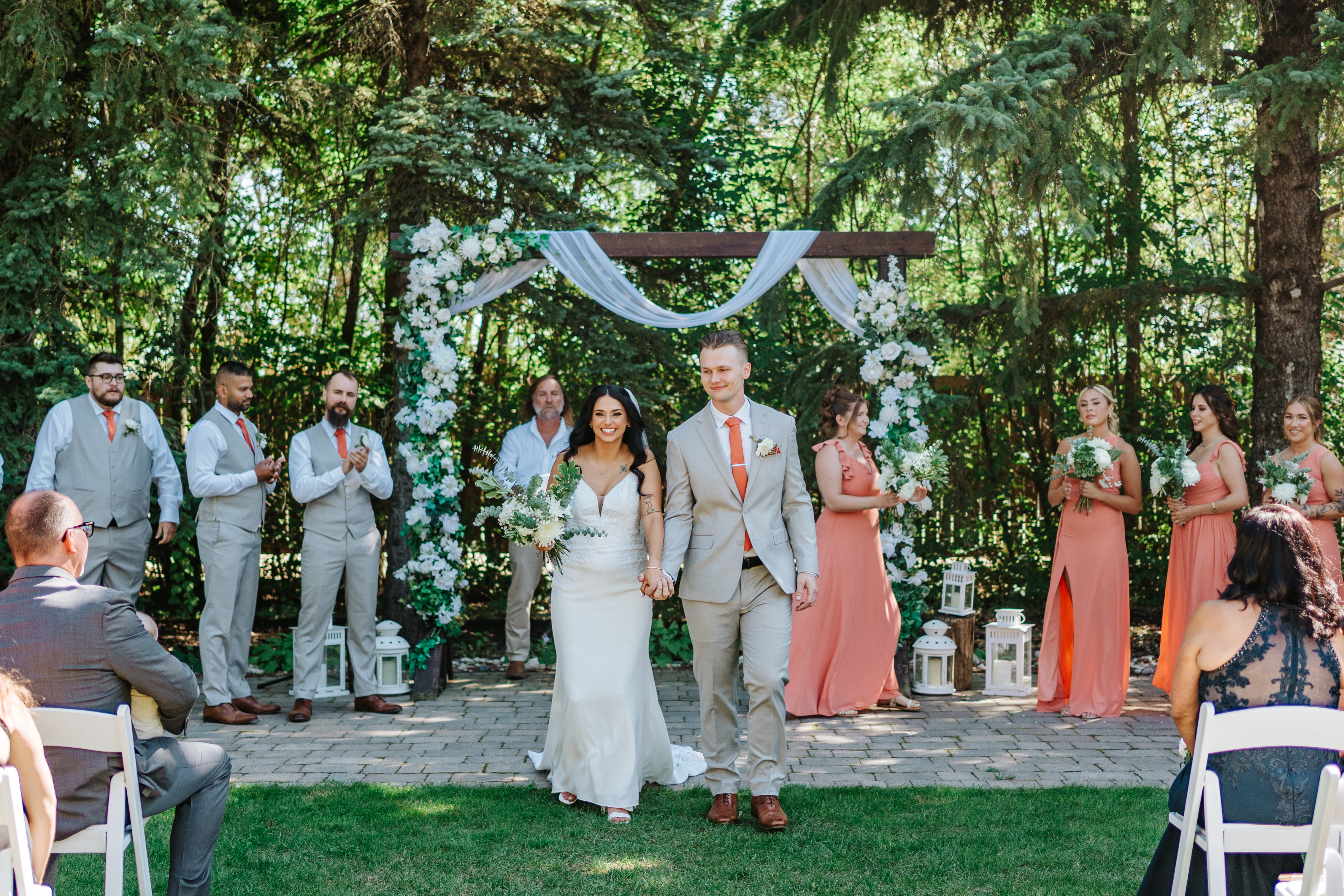 Brandon and Selena at the altar of their outdoor ceremony at Ashgrove Acres with wooden arch, white draping, and coral bridesmaids — Manitoba wedding photography by Chris Ngo