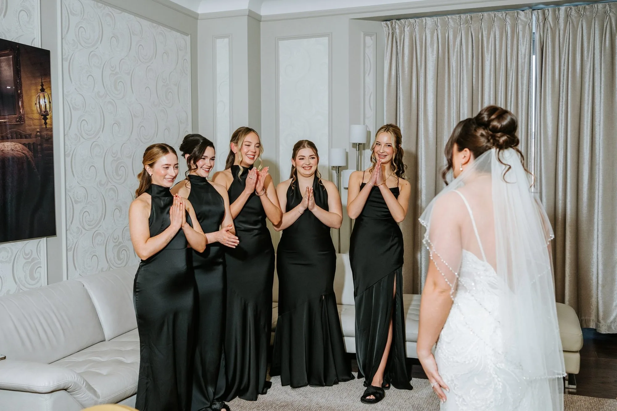 Bridesmaids in black gowns react with joy as the bride reveals her wedding dress — getting-ready moment captured by Winnipeg wedding photographer Ngo Photography