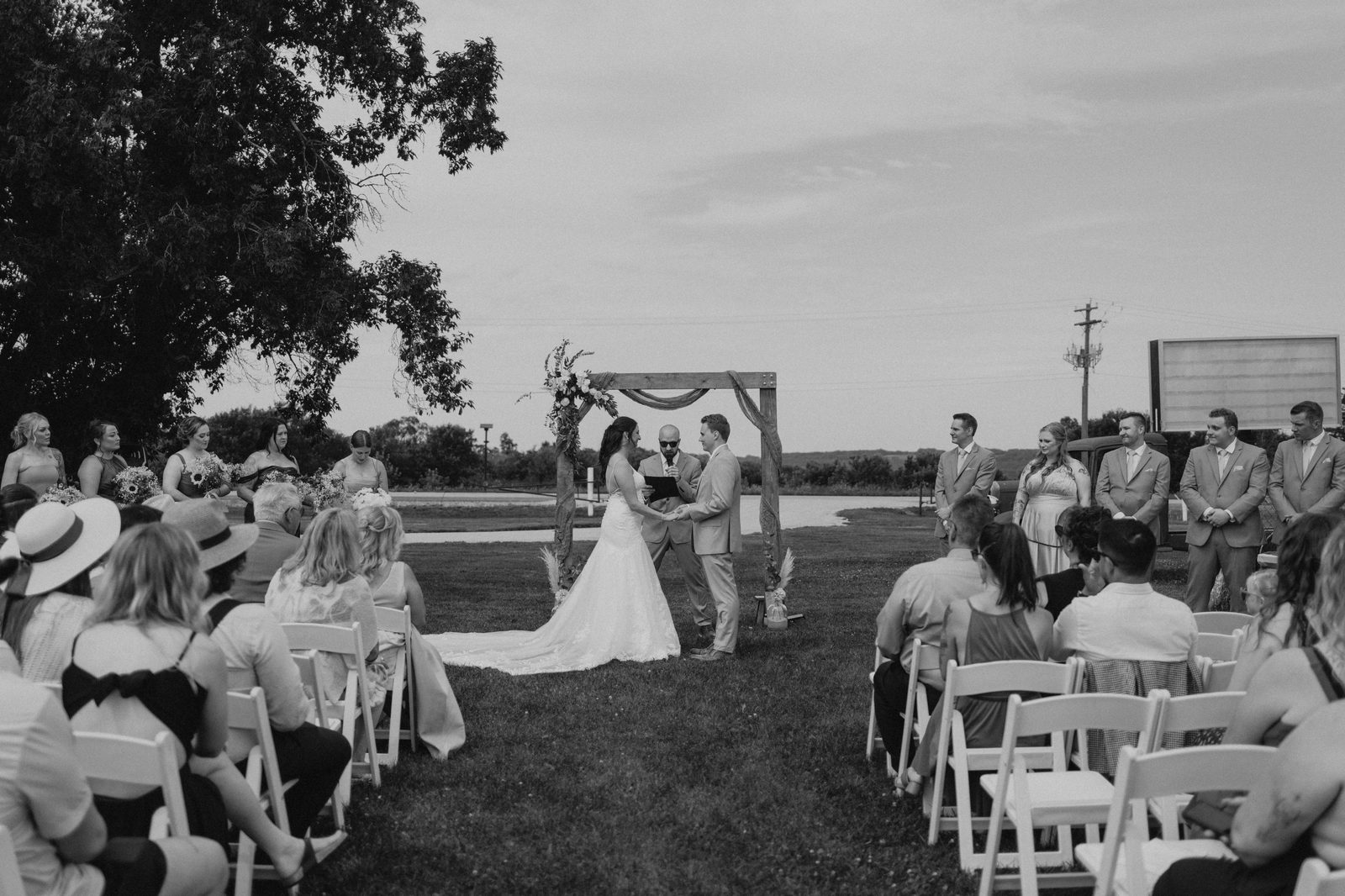 Wide black and white establishing shot of the full ceremony at Pelican Yacht Club — wooden arbor, white chairs, Pelican Lake visible behind — Ninette, Manitoba wedding photography by Chris Ngo