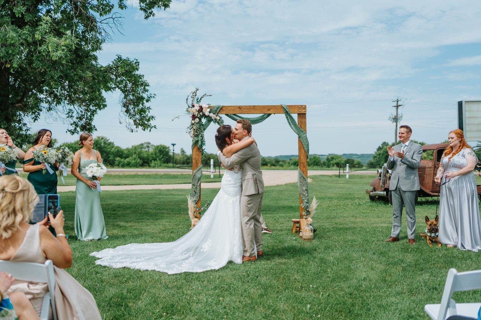 First kiss at the Pelican Yacht Club wooden arbor ceremony — full color wide shot with Pelican Lake in the background — Ninette, Manitoba