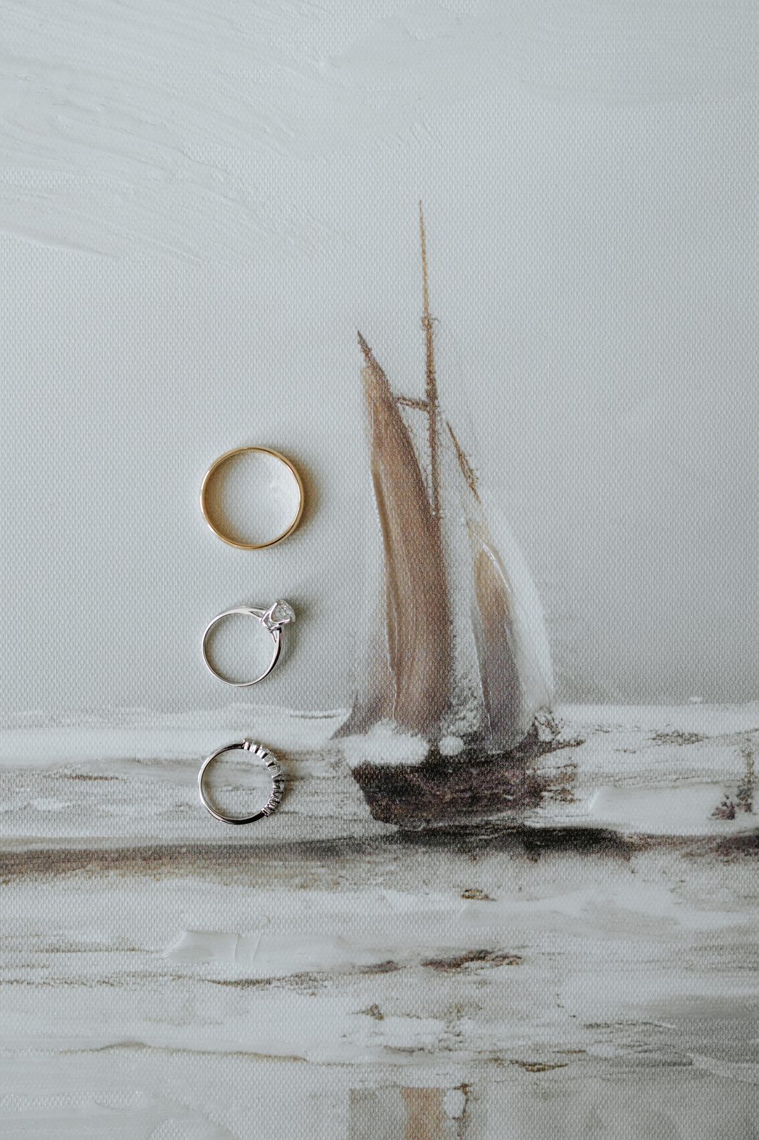 Three wedding rings resting on a sailboat oil painting canvas — heirloom engagement ring, diamond band, and gold wedding band — Pelican Yacht Club wedding, Ninette, Manitoba