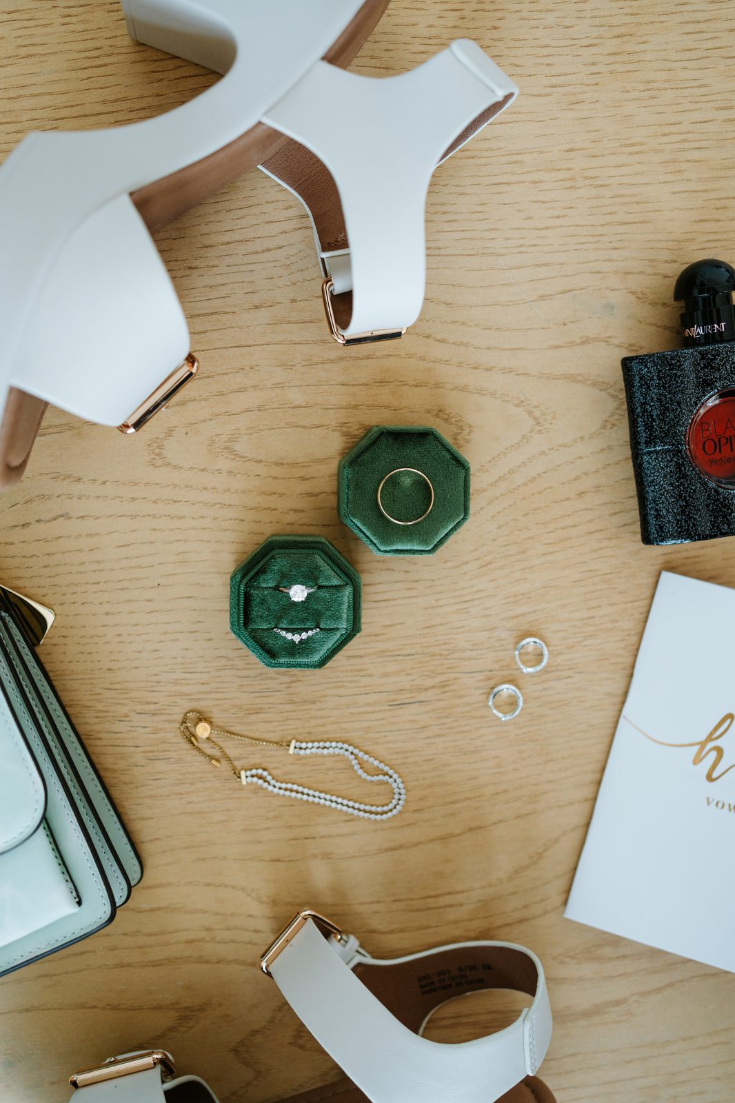 Bridal detail flat lay — green velvet ring boxes with engagement ring, wedding bands, white heels, YSL Black Opium perfume, pearl necklace, and vow book — Pelican Yacht Club wedding, Ninette, Manitoba