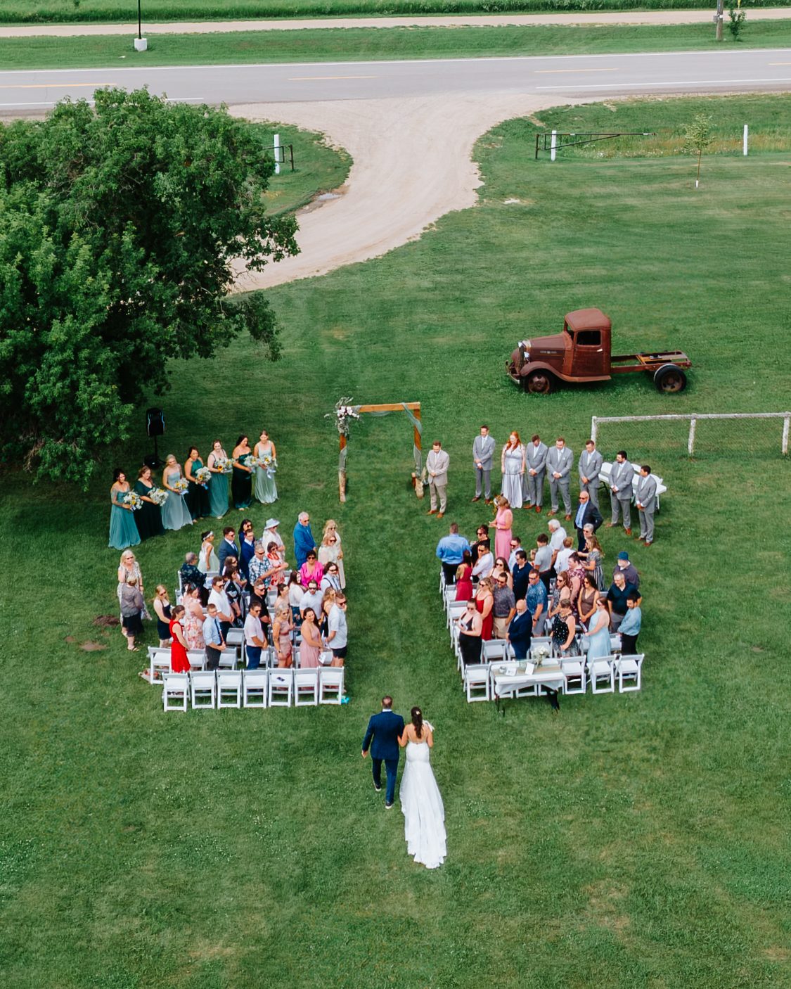 Aerial drone shot of the wedding ceremony at Pelican Yacht Club — bride walking down the aisle with the full ceremony visible from above, Pelican Lake behind — Ninette, Manitoba
