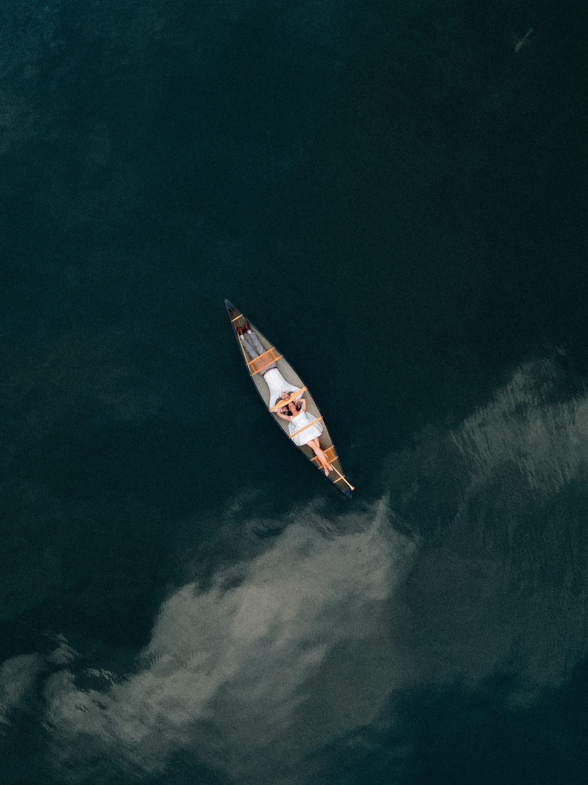 Black and white aerial drone — bride Christina in the canoe on dark Pelican Lake, wedding dress visible — Ninette, Manitoba