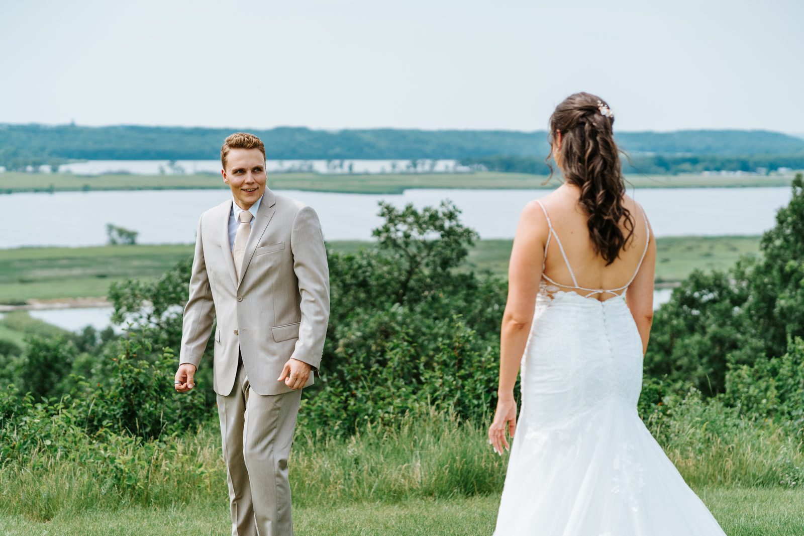 First look on the hillside above Pelican Lake — groom turning to see bride, Pelican Lake spanning the full horizon behind her — Pelican Yacht Club, Ninette, Manitoba