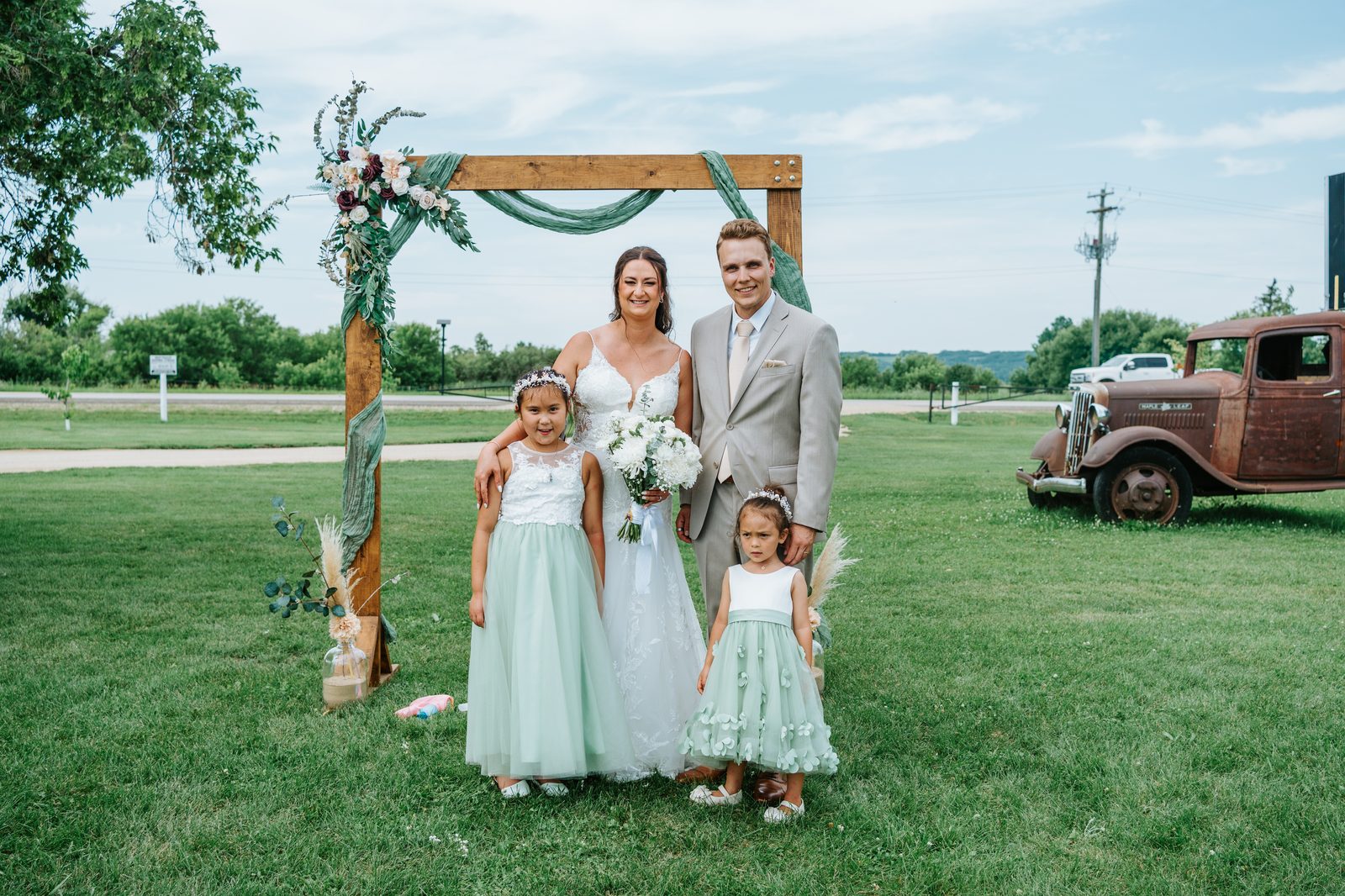 Christina and Jack with their two flower girls in mint green dresses at the wooden arbor — Pelican Yacht Club wedding, Ninette, Manitoba