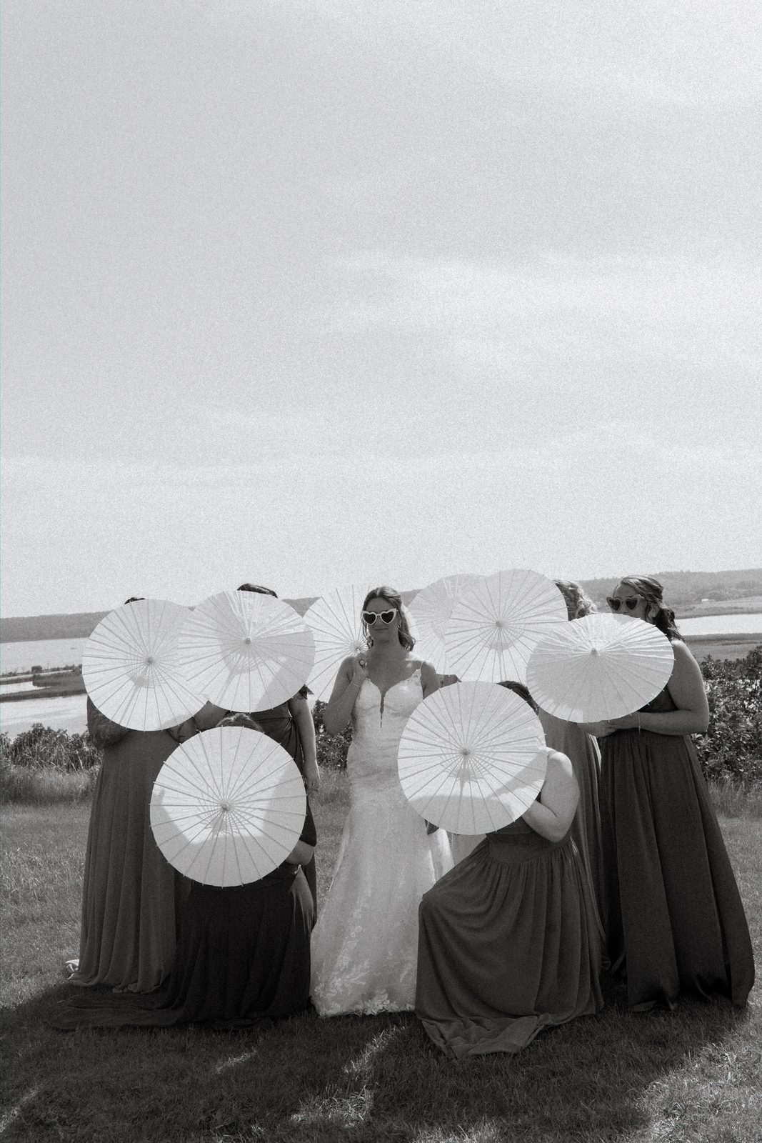 Black and white parasol portrait — bridesmaids hiding behind parasols, only Christina visible with lake behind her — Pelican Yacht Club, Ninette, Manitoba