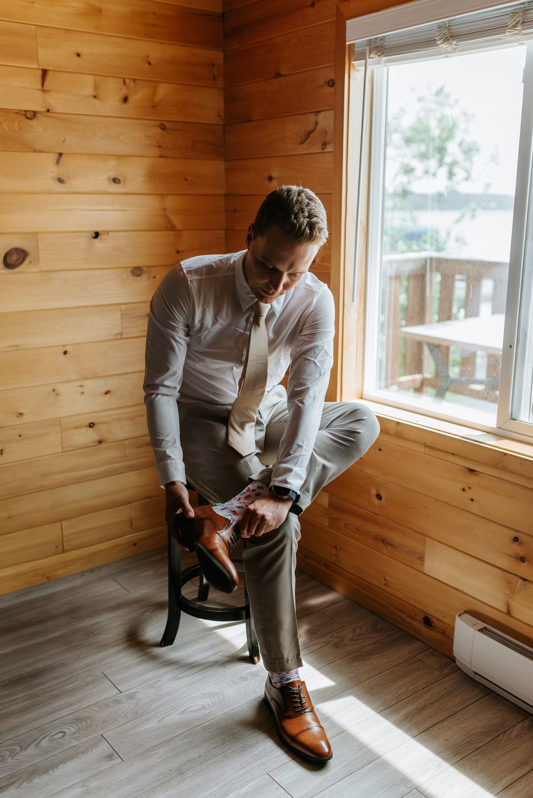 Groom Jack tying his brown Oxford shoes in the knotty pine cabin at Pelican Yacht Club, lake visible through the window — Ninette, Manitoba wedding photographer Chris Ngo