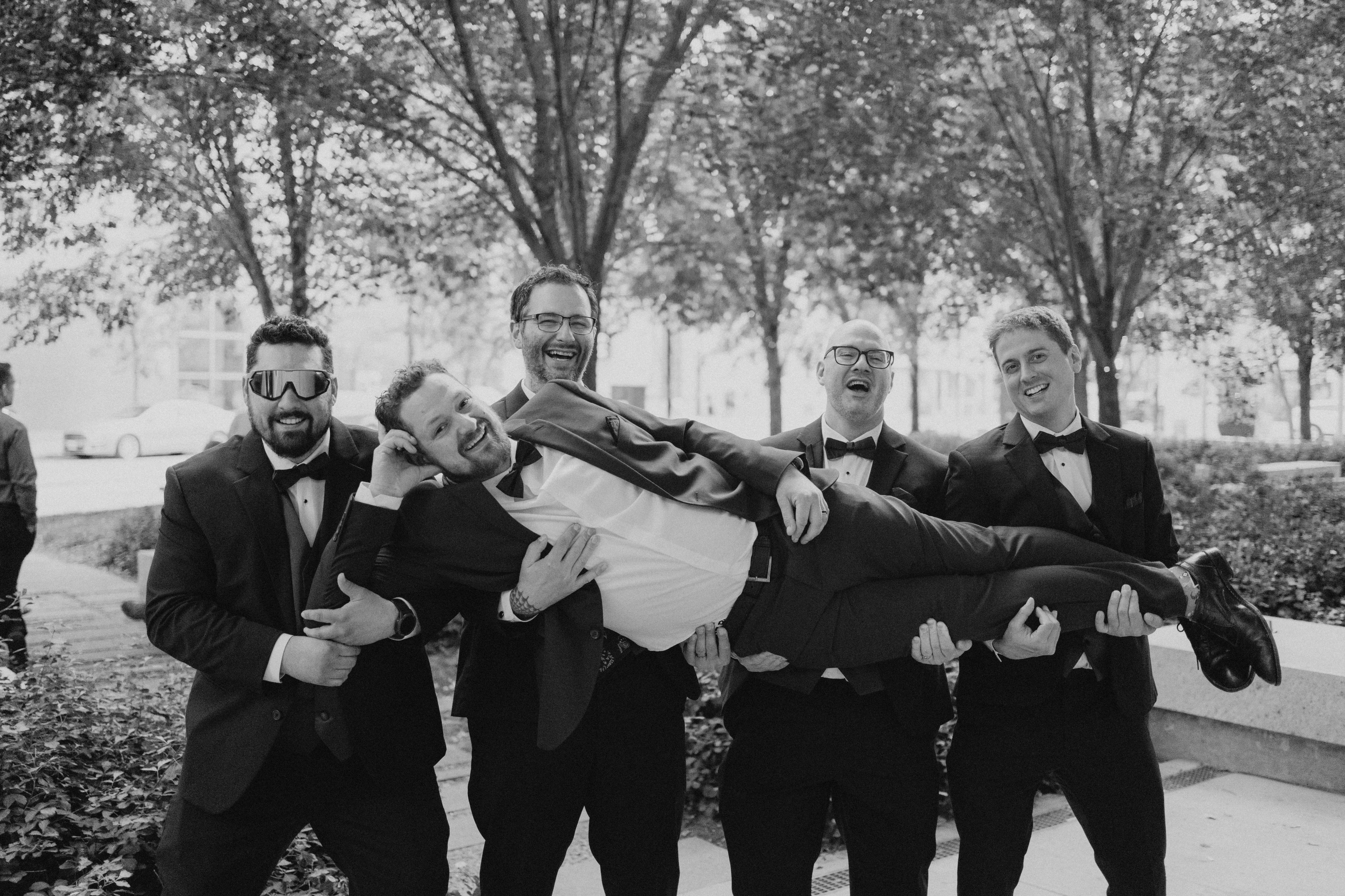 Dave's groomsmen lifting him off the ground in tuxedos outside their hotel in downtown Winnipeg — wedding photography by Chris Ngo