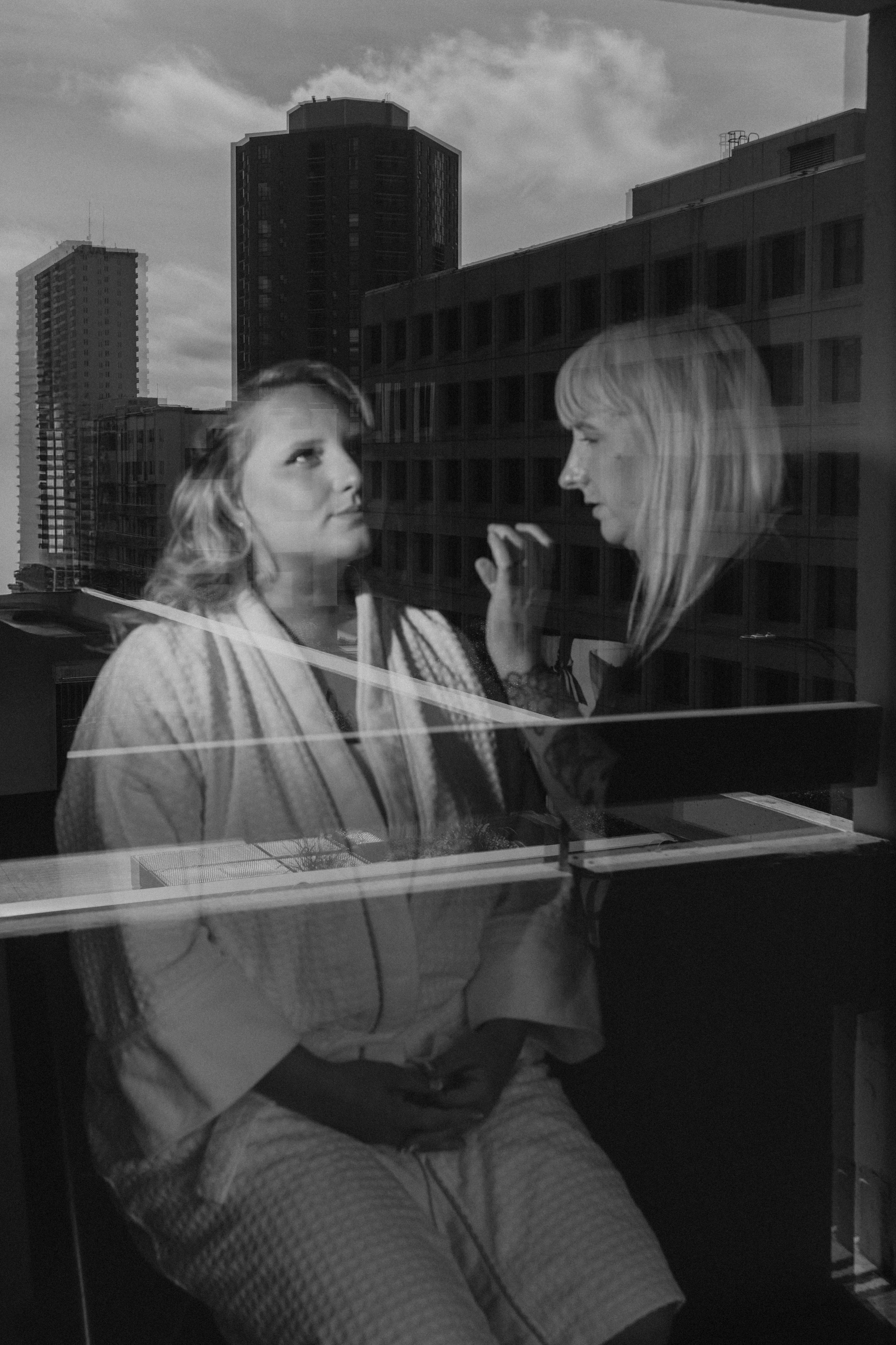 Bailey getting ready in her hotel room, seen through the window glass with the Winnipeg downtown skyline reflected behind her — wedding photography by Chris Ngo