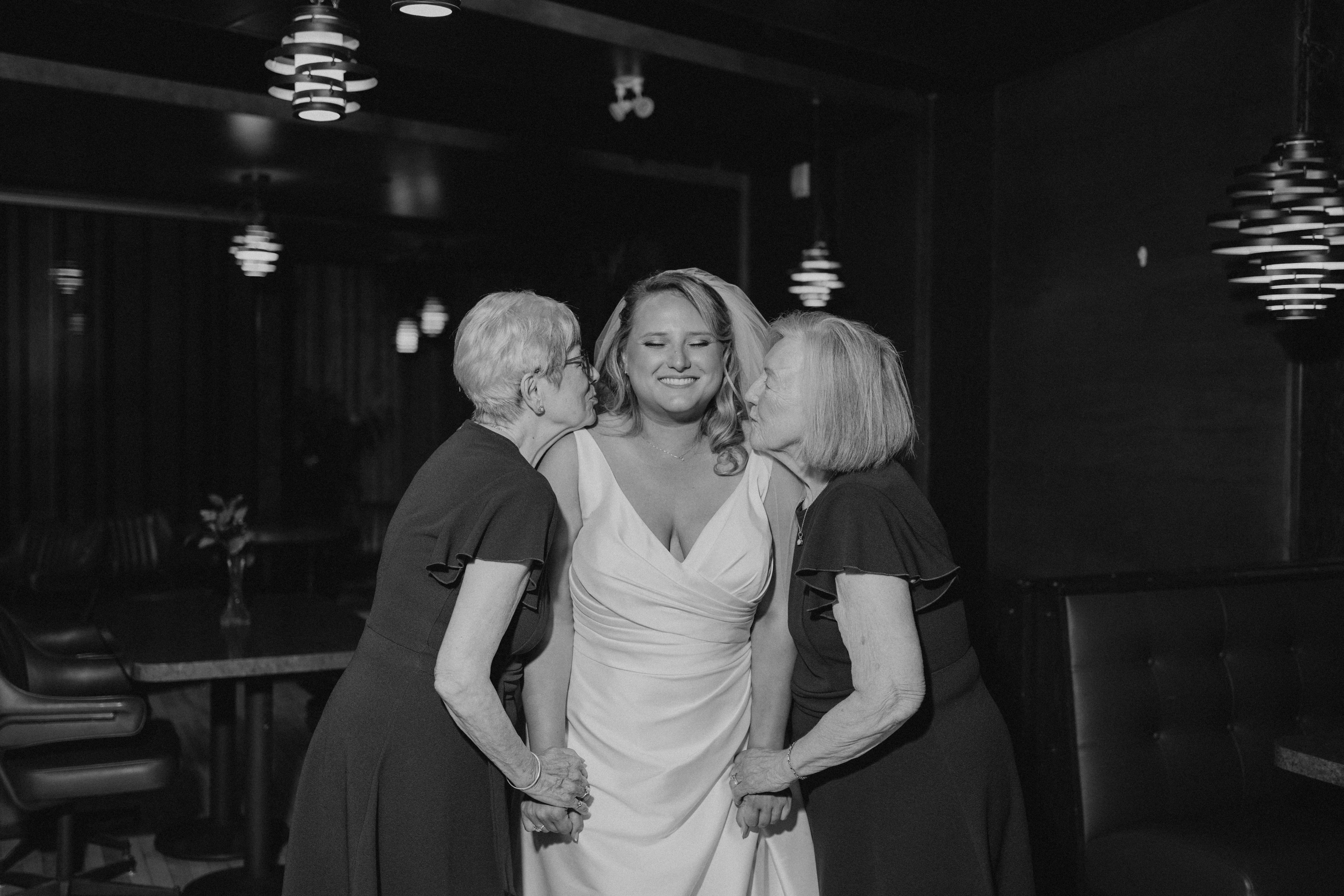 Bailey's two grandmothers leaning in to kiss her cheeks simultaneously at the reception inside Rudy's — Winnipeg wedding photography by Chris Ngo