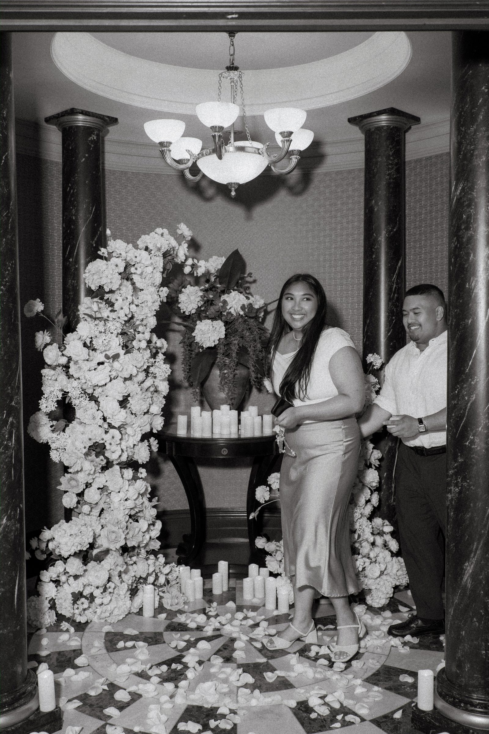 Wide shot of Deril and Annie framed by the twin floral arches with chandelier above and rose petals below — surprise proposal photography downtown Winnipeg by Ngo Photography