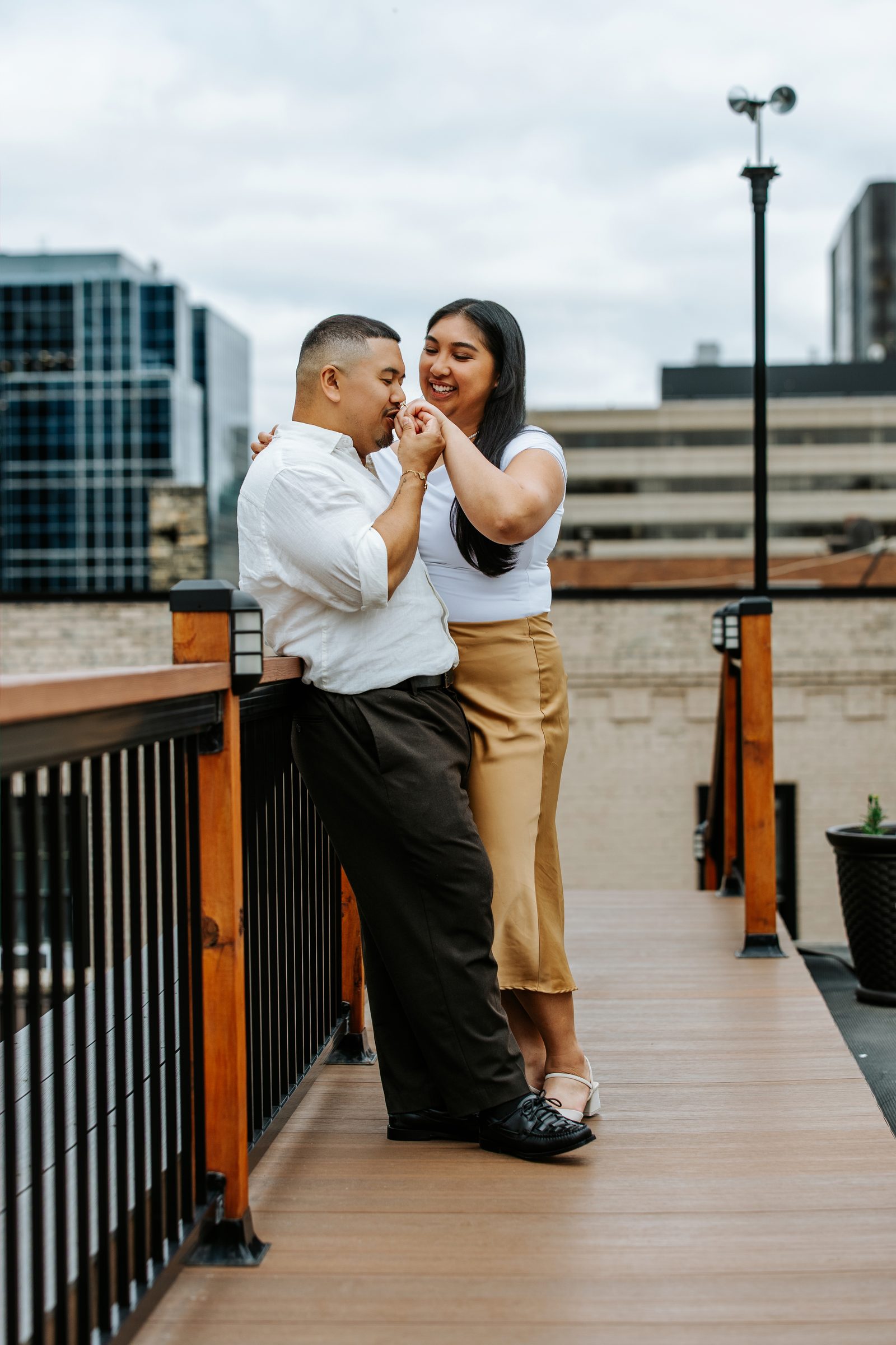 Annie and Deril candid moment on the rooftop with the downtown Winnipeg cityscape behind them — just engaged — Ngo Photography