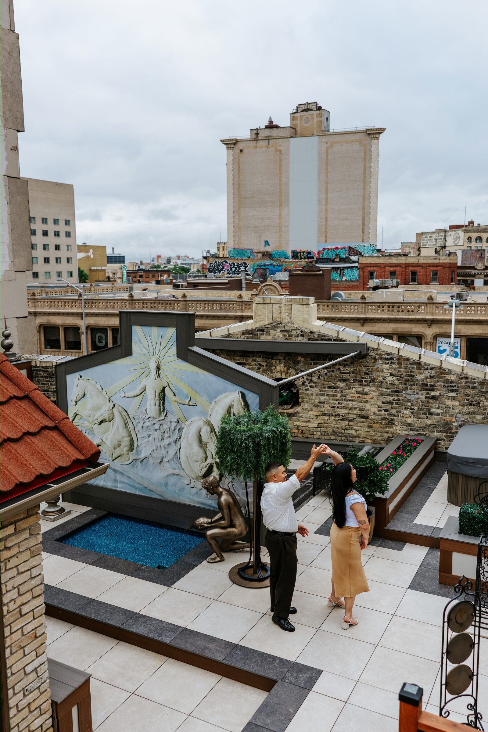 Annie and Deril on the rooftop steps with the downtown Winnipeg skyline and classical stone buildings — engagement portraits by Ngo Photography