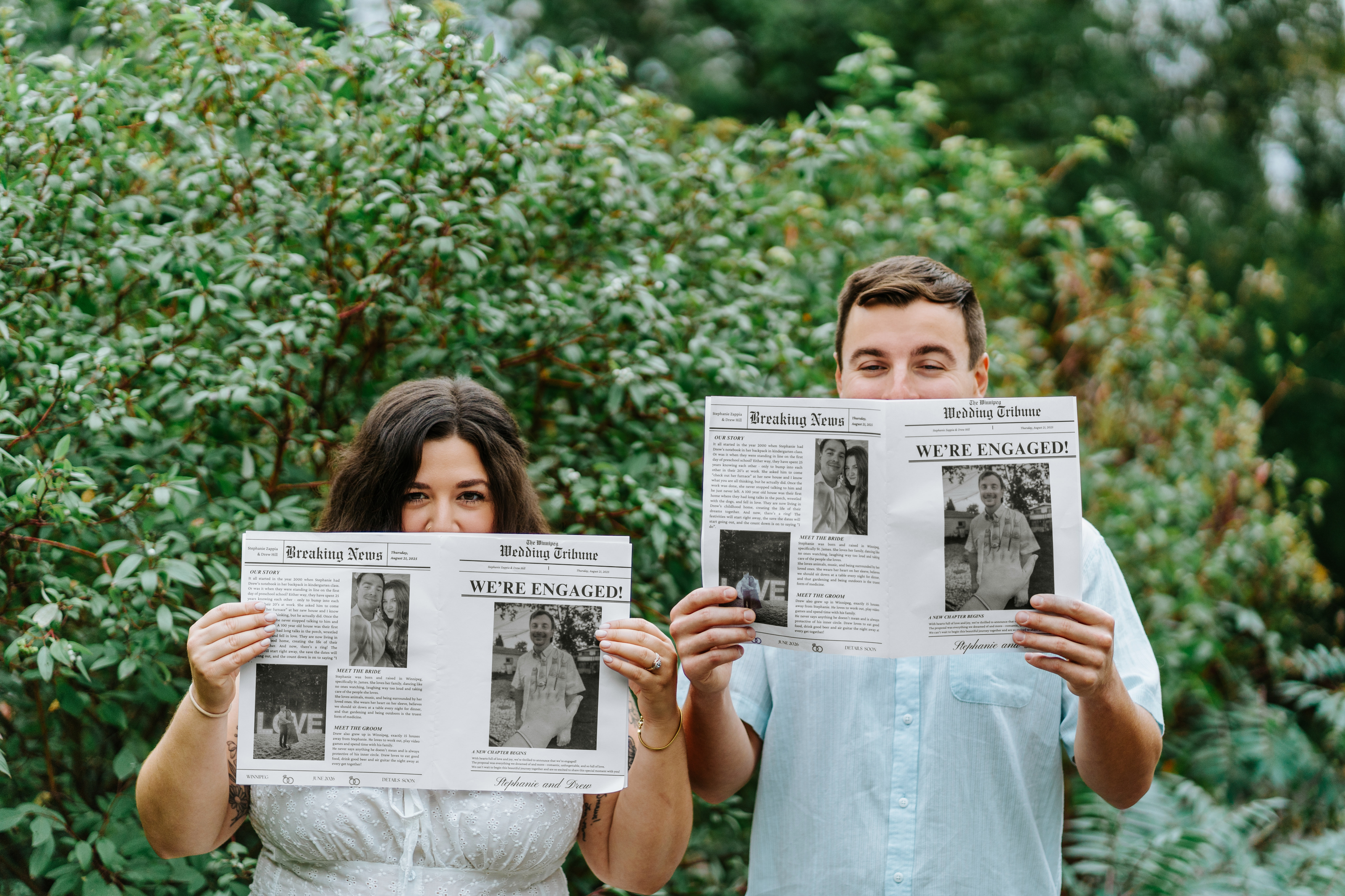 Drew and Stephanie holding We're Engaged newspapers in front of the lush garden greenery at Assiniboine Park Winnipeg — engagement photos by Ngo Photography