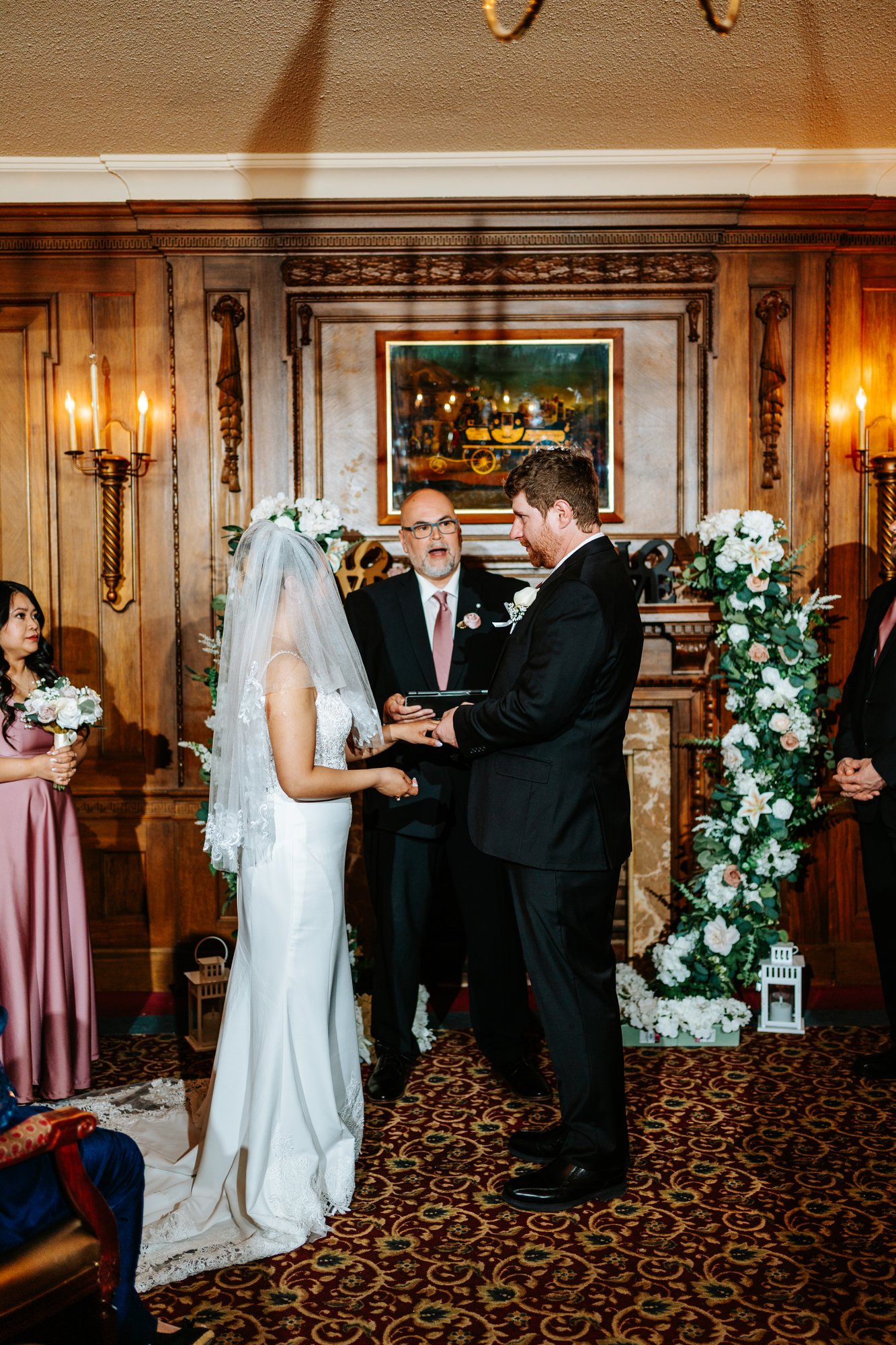 Couple exchanging vows at the carved Victorian fireplace with white floral columns at Bailey's Restaurant Winnipeg — wedding ceremony photography by Ngo Photography