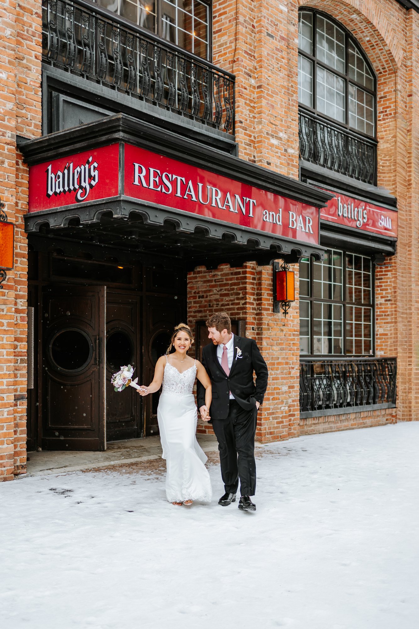 Eden and Justin outside Bailey's Restaurant and Bar on Bannatyne Avenue Winnipeg, snow on the ground, red sign and brick building behind them — wedding photography by Ngo Photography