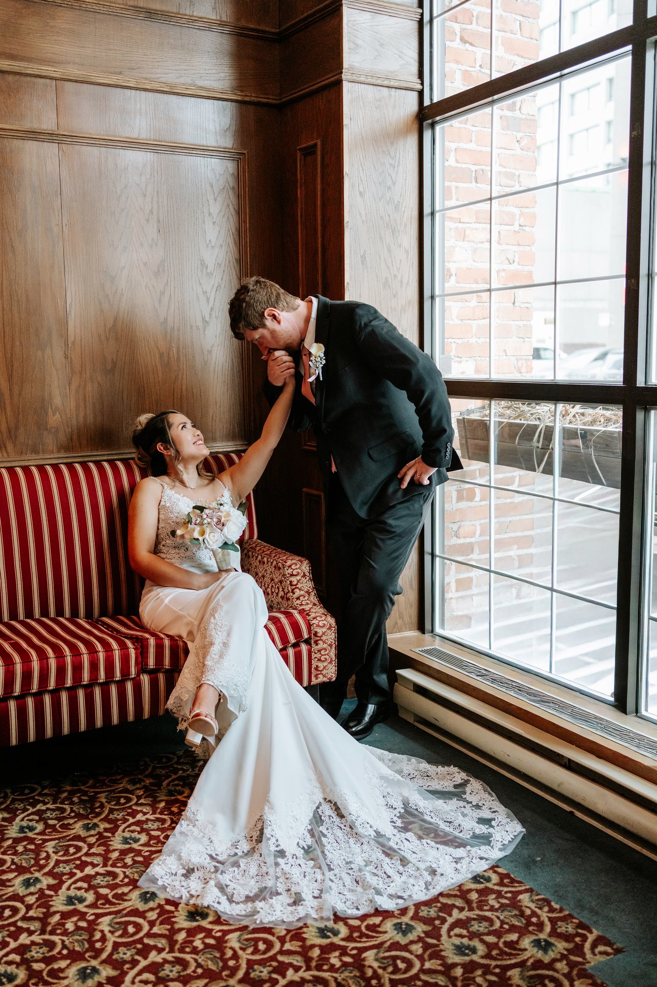 Justin kissing Eden's hand as she looks up at him from the red sofa at Bailey's Restaurant Winnipeg — candid wedding portrait by Ngo Photography