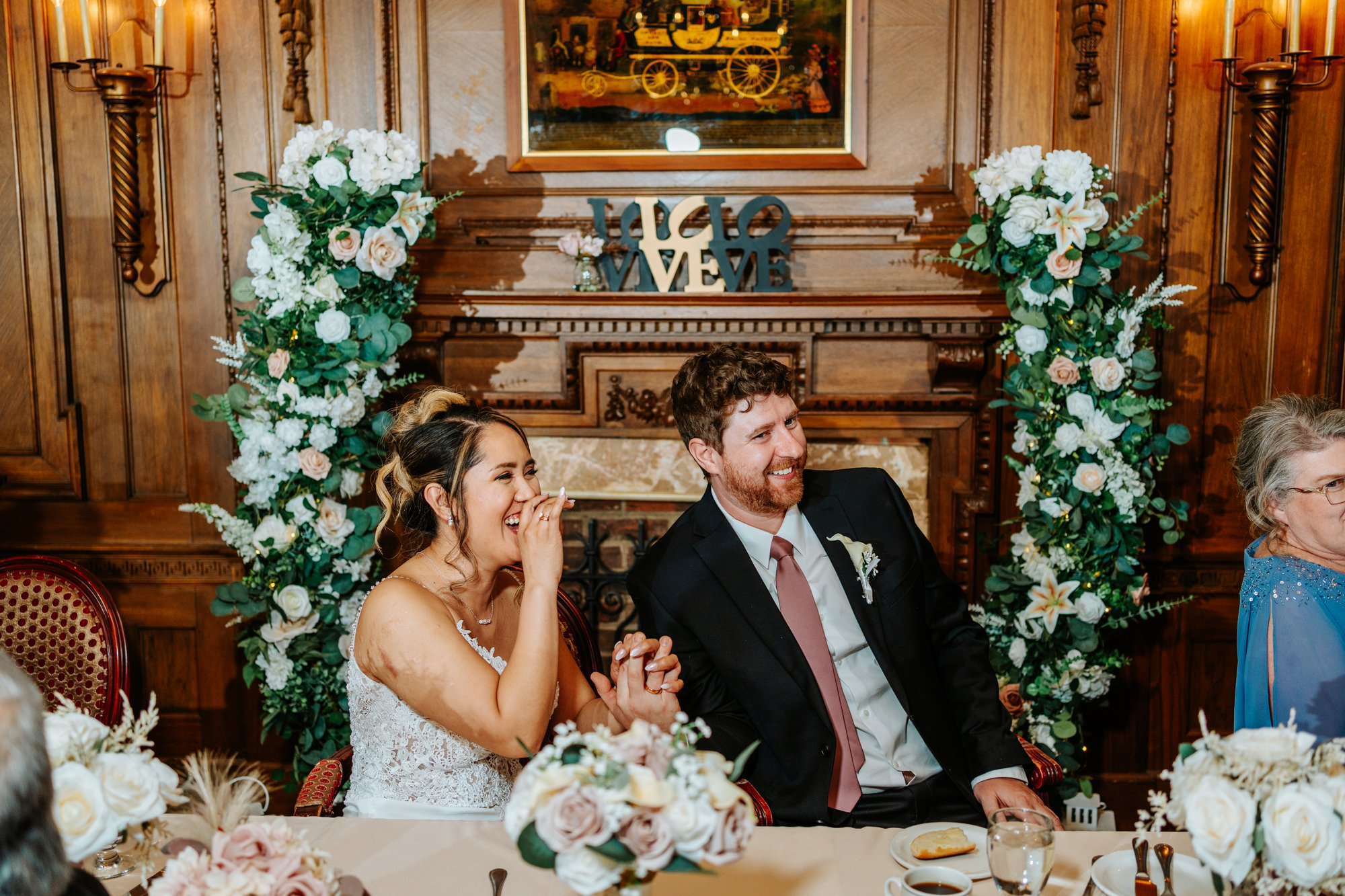 Eden and Justin laughing at the head table at Bailey's Restaurant wedding reception, white floral arrangements flanking the carved fireplace — Ngo Photography Winnipeg