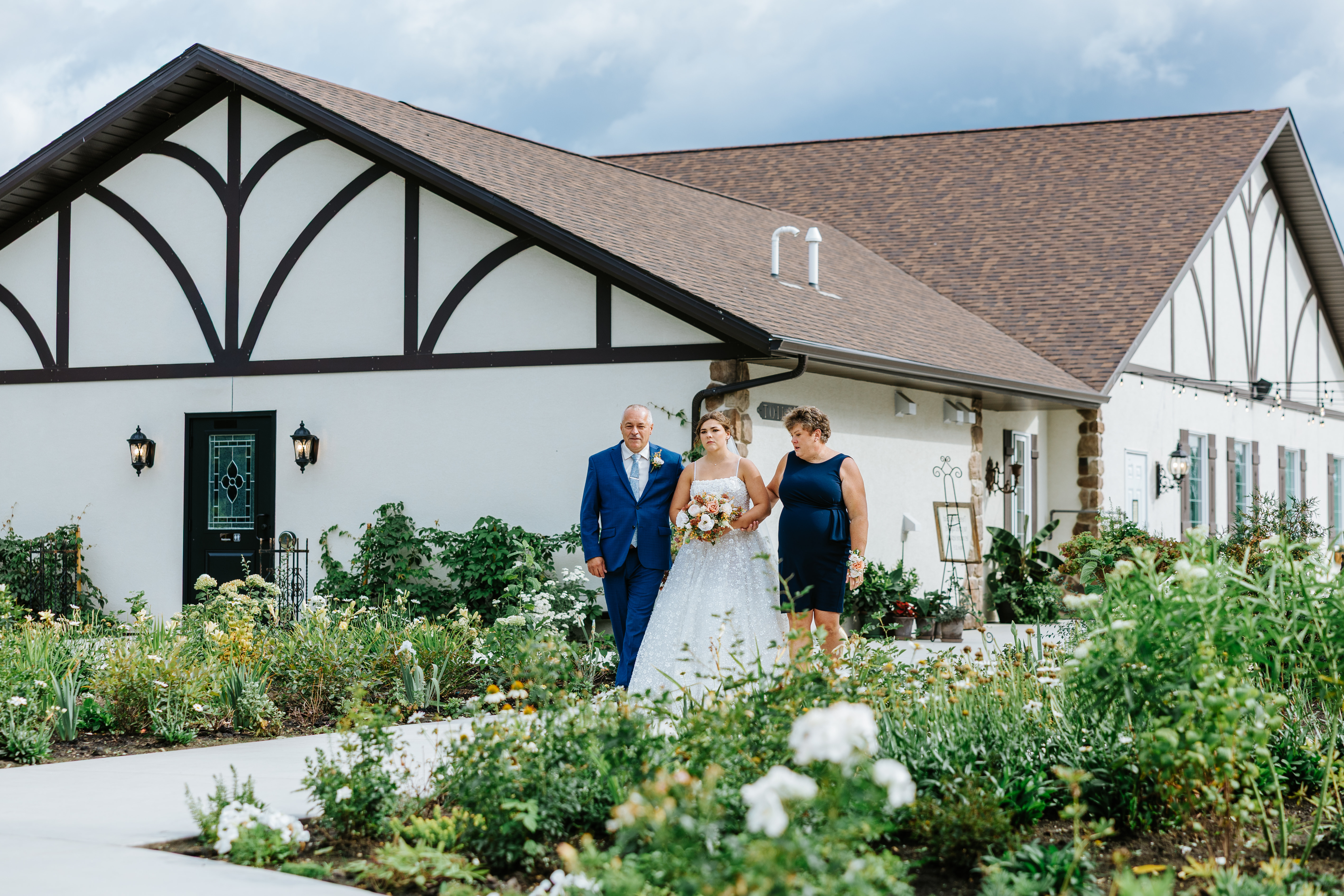 Erin walking down the garden path toward the pavilion ceremony flanked by both parents at Buttercup Café Manitoba — processional wedding photography by Ngo Photography