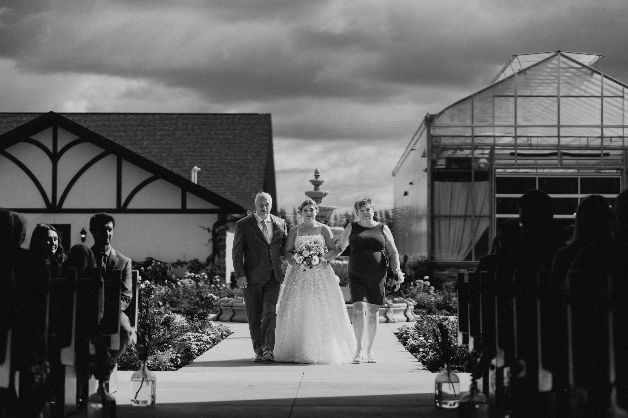Erin walking down the outdoor ceremony aisle at Buttercup Café Manitoba flanked by her parents — black and white wedding photography by Ngo Photography