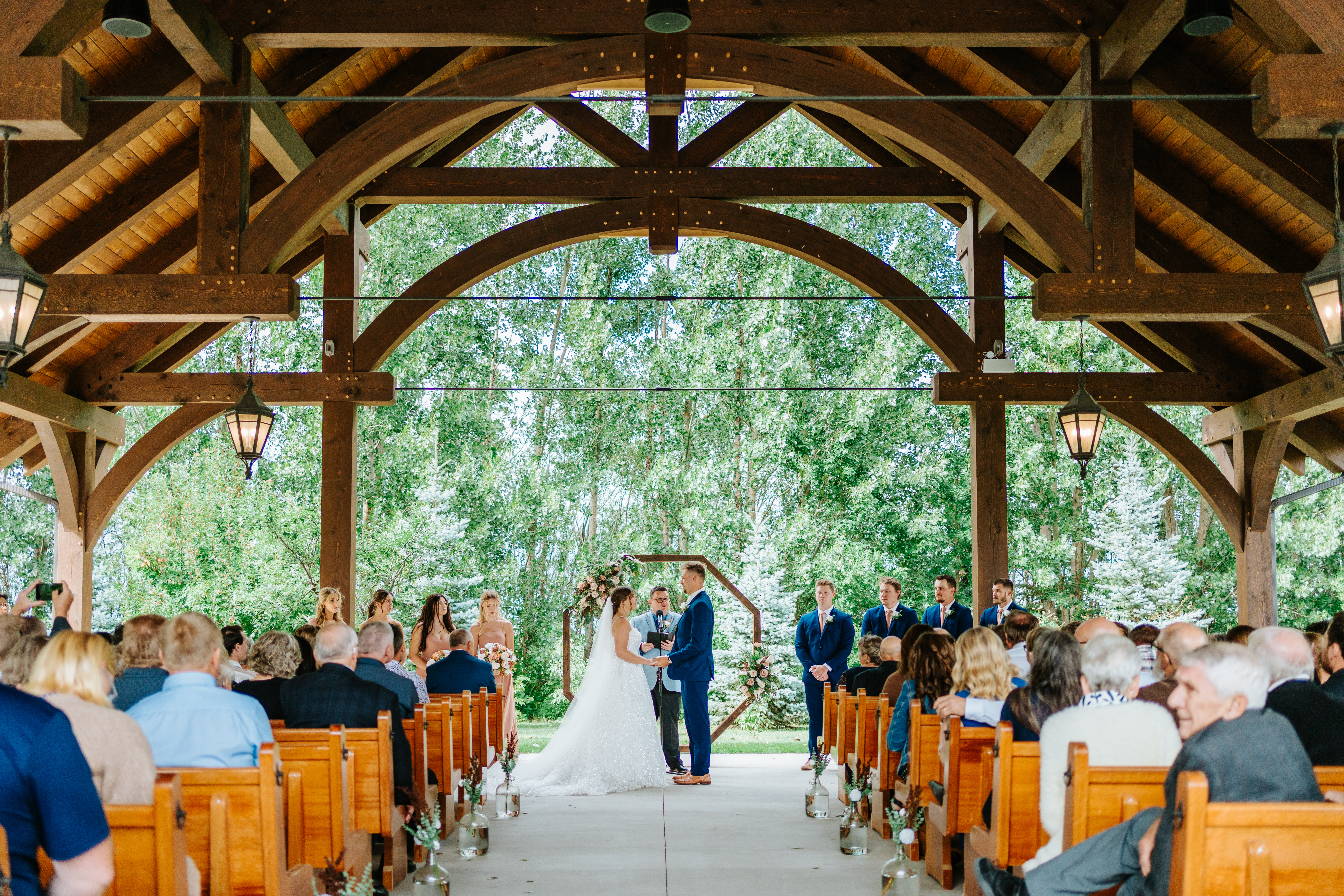 Wide view of Erin and Jayden's outdoor ceremony in the timber pavilion at Buttercup Café Manitoba — all guests seated, couple at the hexagonal arch — wedding photography by Chris Ngo