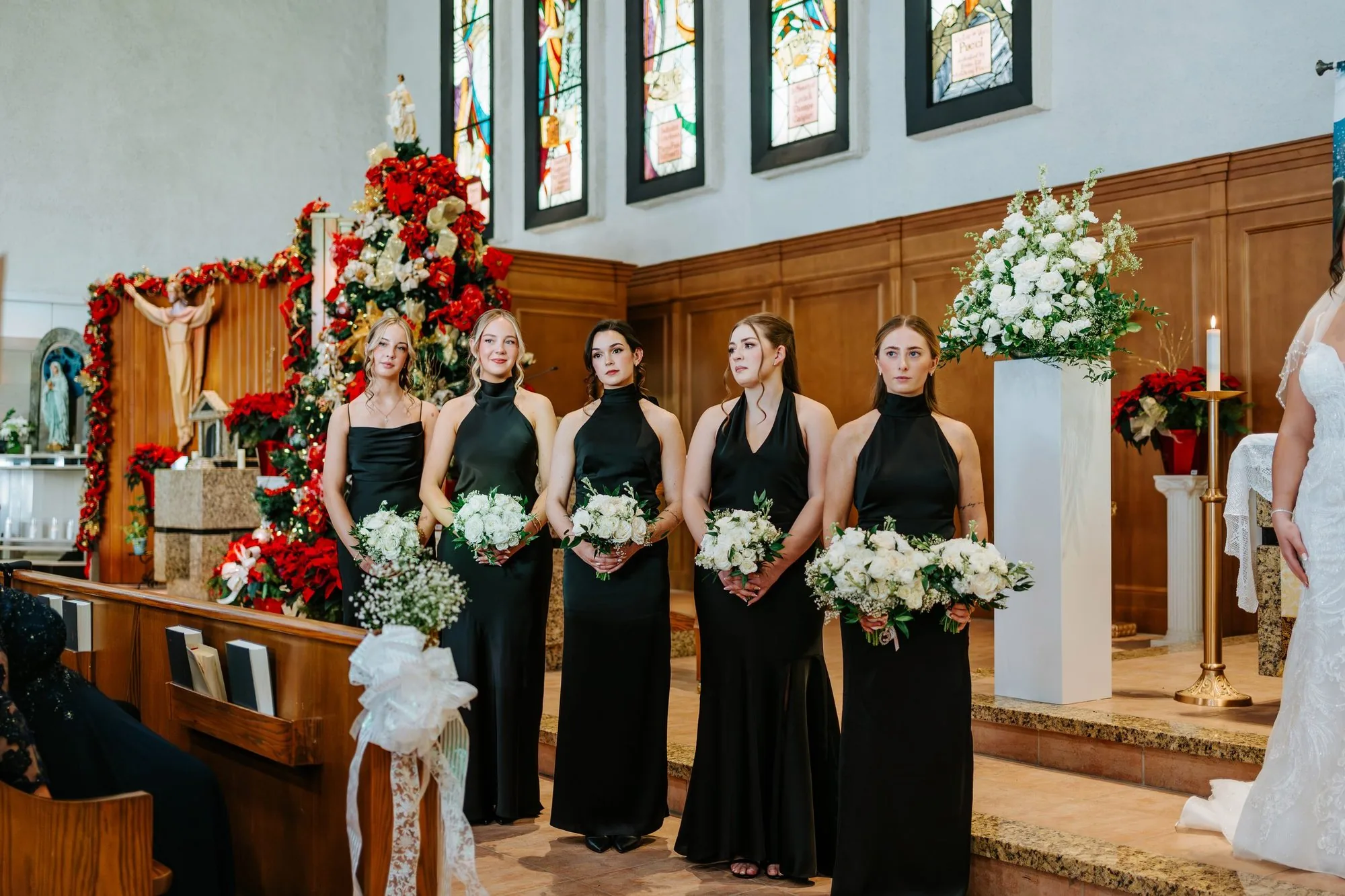 Bridesmaids in black dresses holding white bouquets inside a church with stained glass windows — Winnipeg wedding photography by Ngo Photography