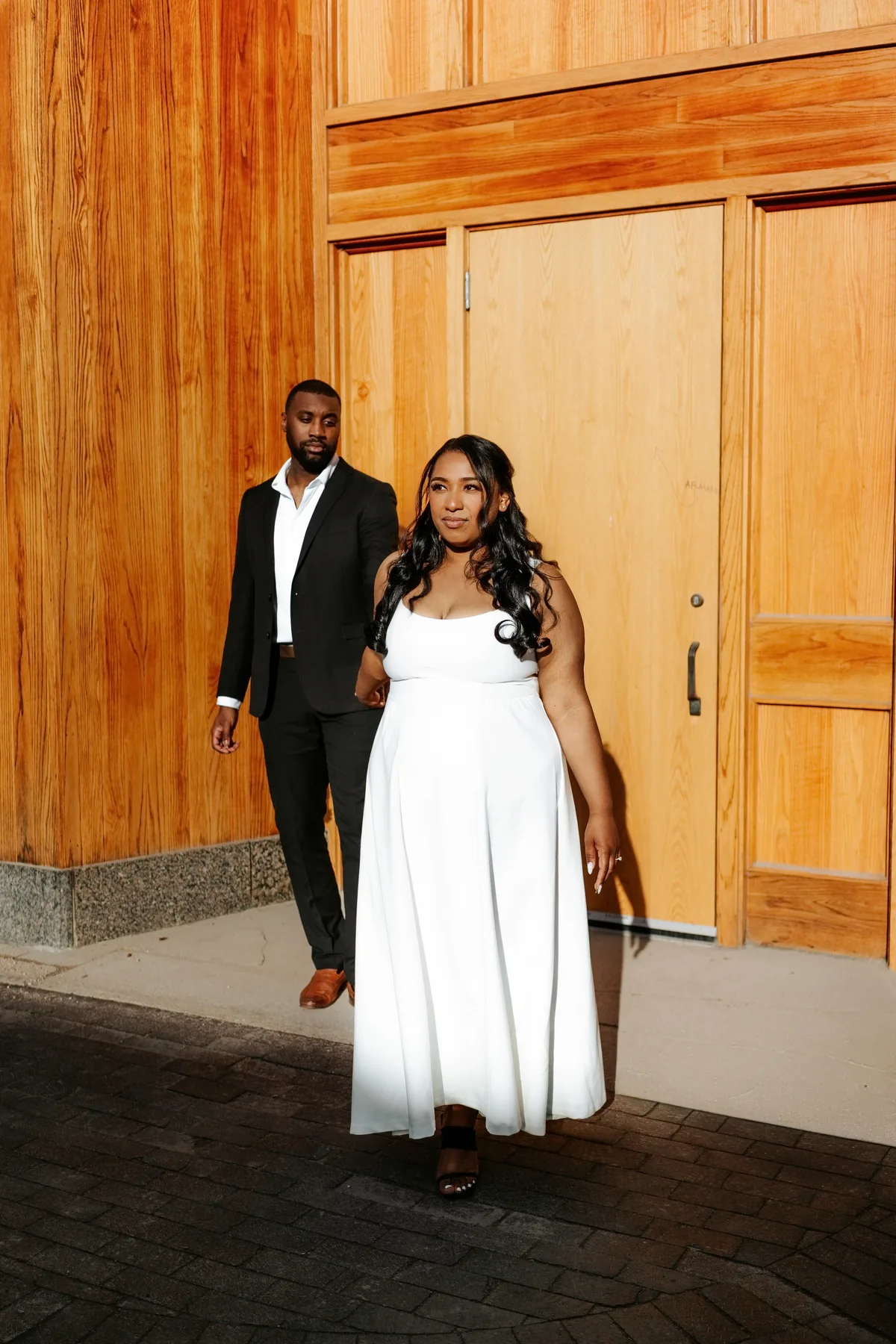 Jaida and Jordan walking together in front of warm wooden doors at the CMHR, downtown Winnipeg wedding — Ngo Photography