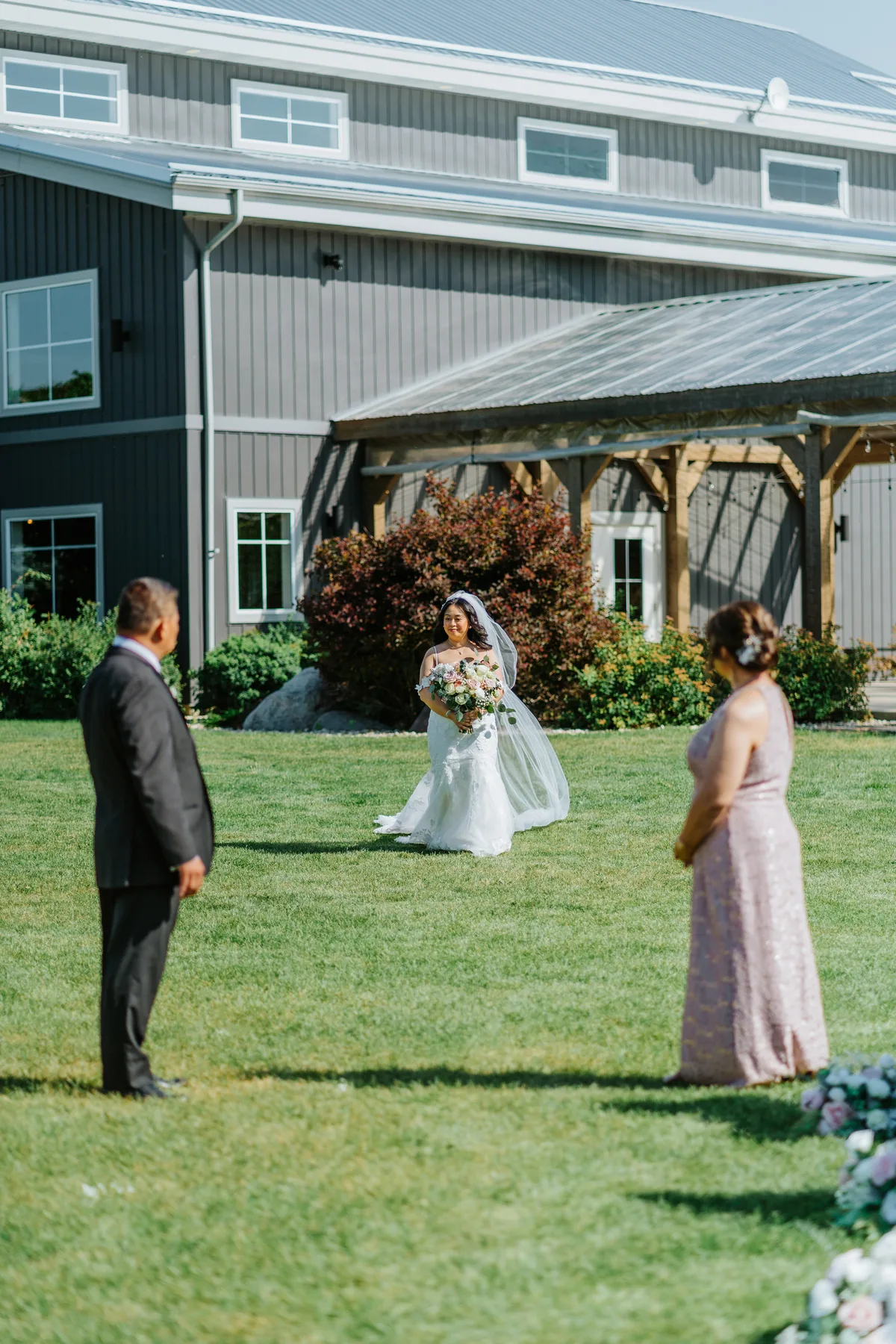 Jay walking down the aisle with her parents, veil flowing, toward the arbor at Hawthorne Estates, Manitoba