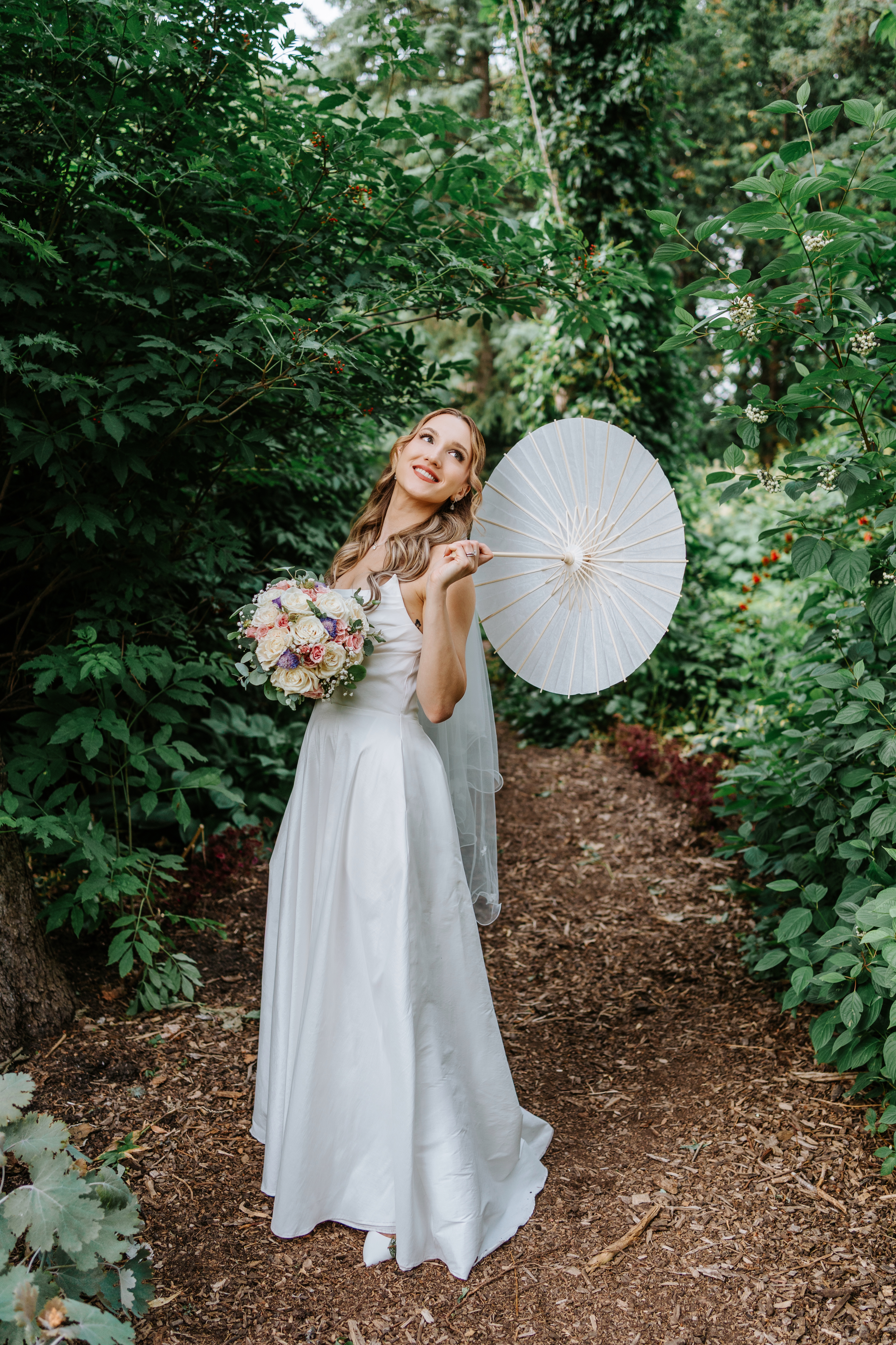 Tanya in white wedding gown holding a parasol and colorful bouquet in Assiniboine Park garden — Ngo Photography Winnipeg