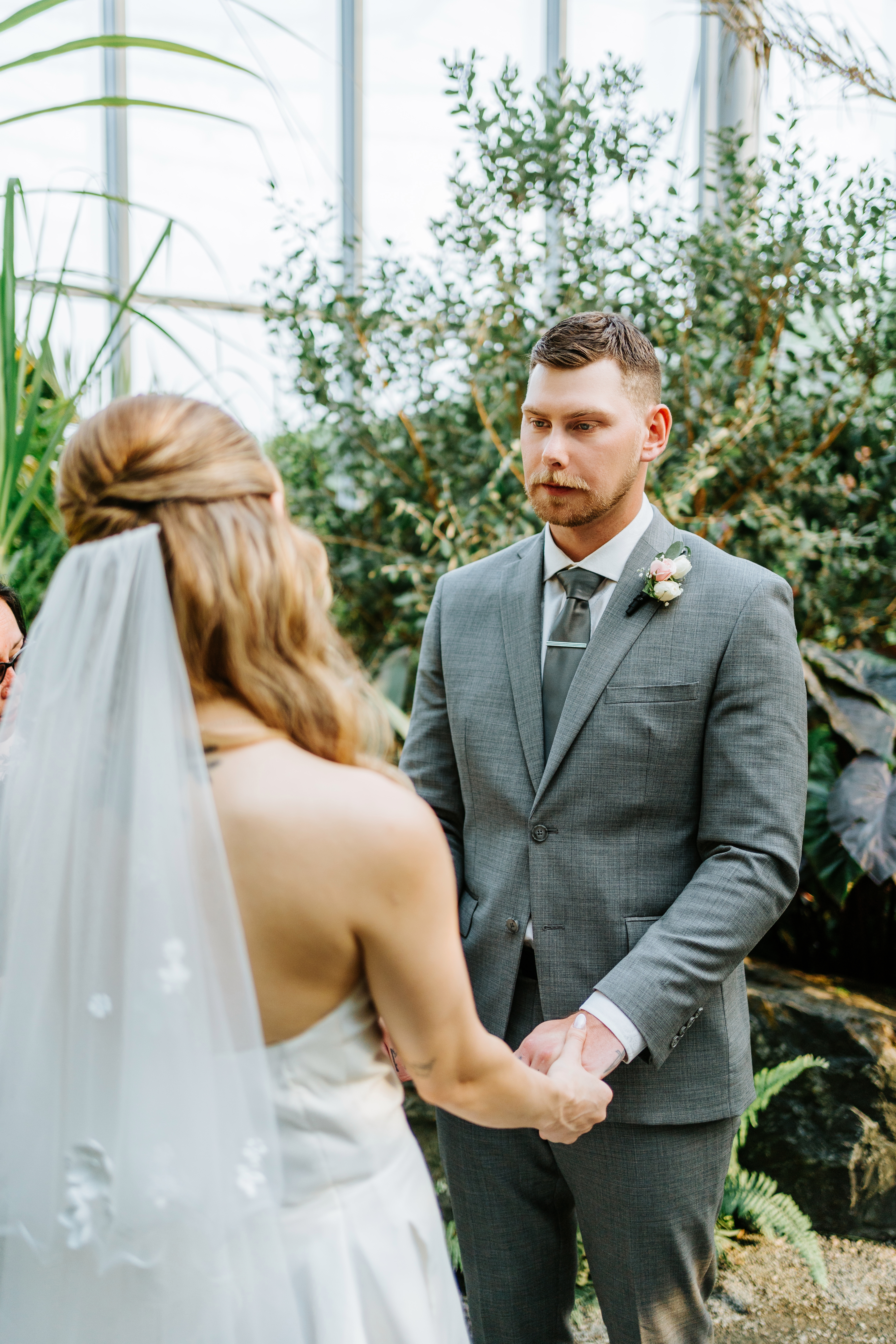 Liam holding Tanya's hands during their intimate wedding ceremony inside The Leaf at Assiniboine Park, lush tropical plants and glass ceiling behind — wedding photography by Chris Ngo