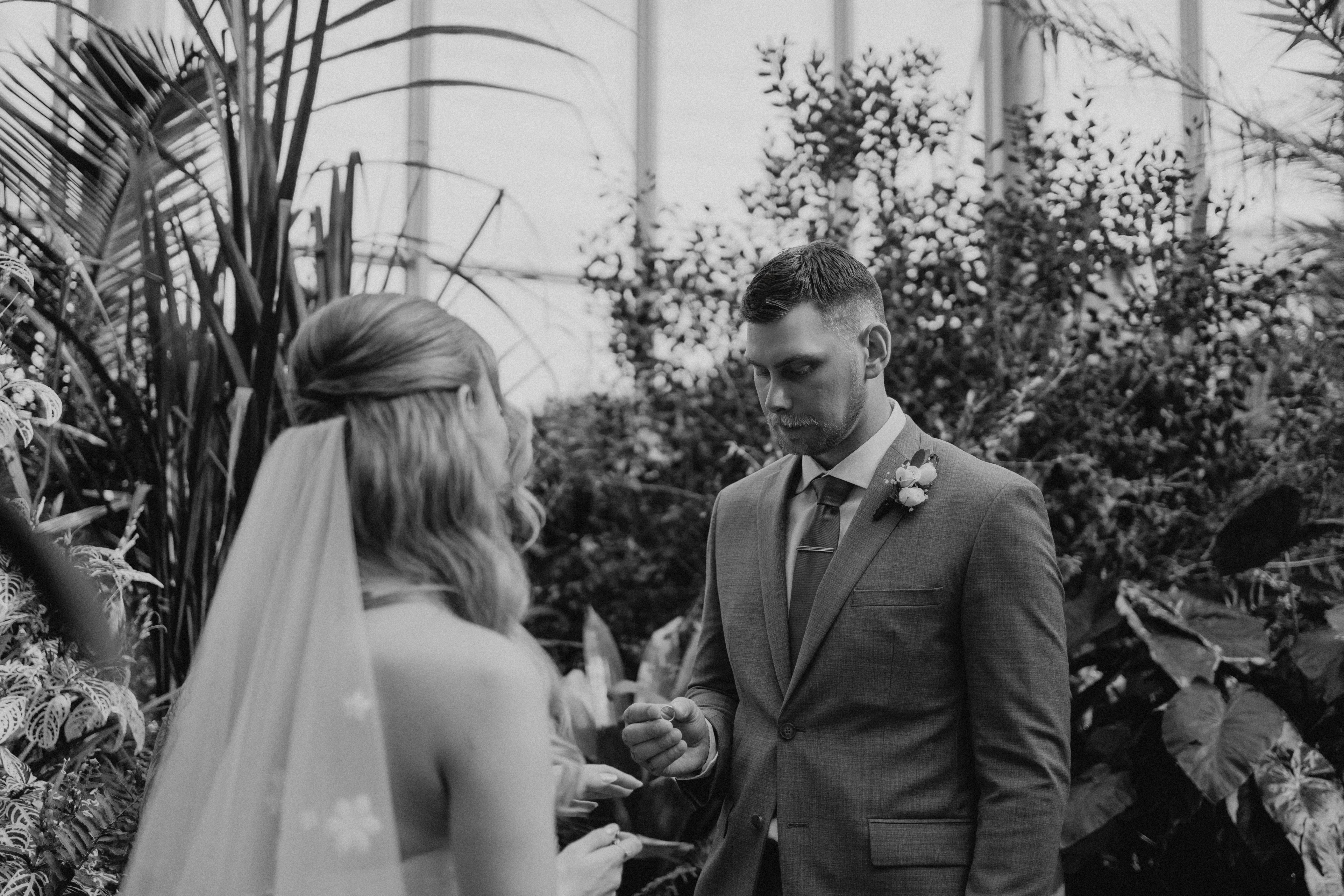 Black and white photo of Liam placing the ring on Tanya's finger during their intimate ceremony inside The Leaf, tropical palms behind — Ngo Photography Winnipeg