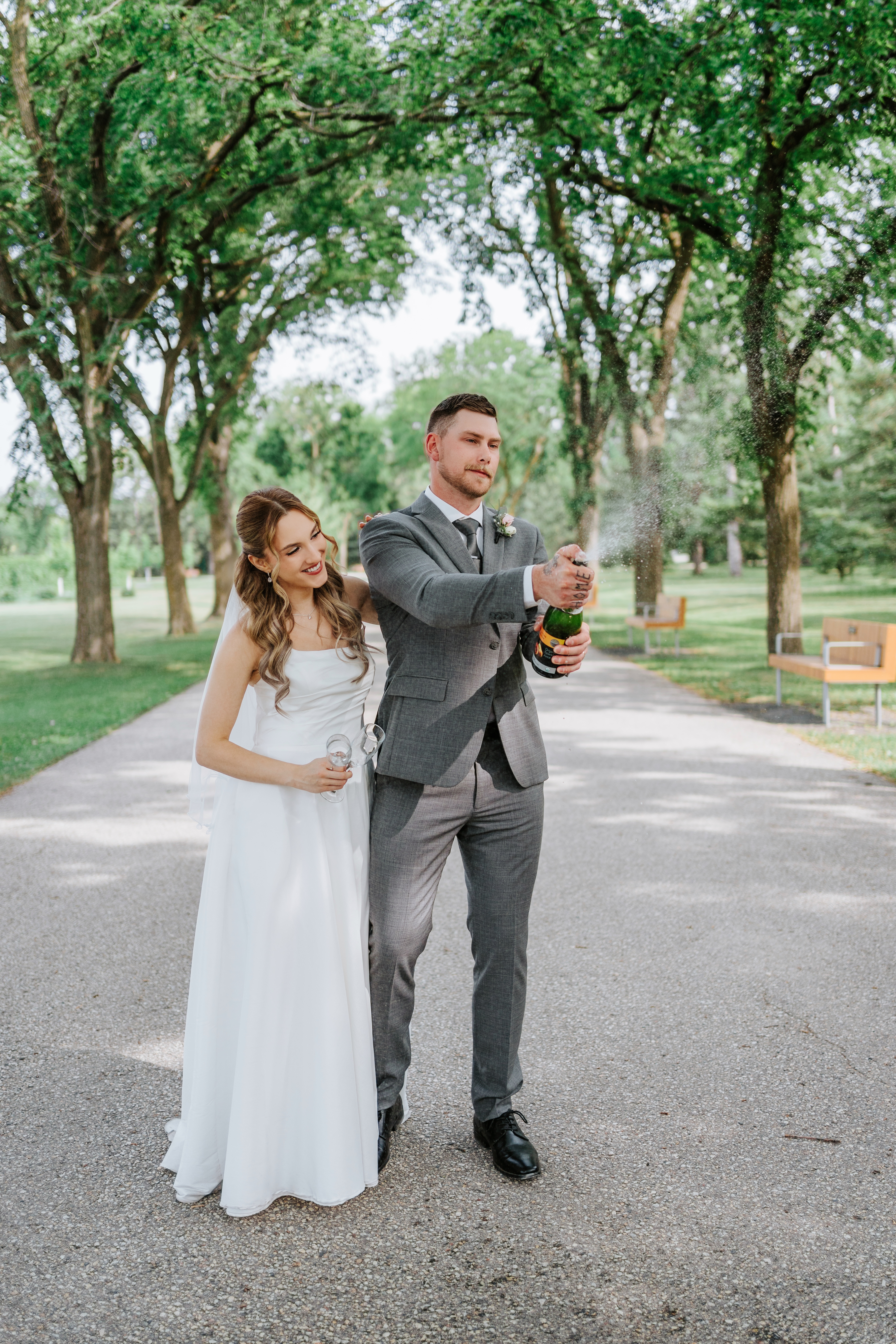 Liam popping champagne bottle on the Assiniboine Park promenade with Tanya laughing beside him — wedding celebration photography by Chris Ngo