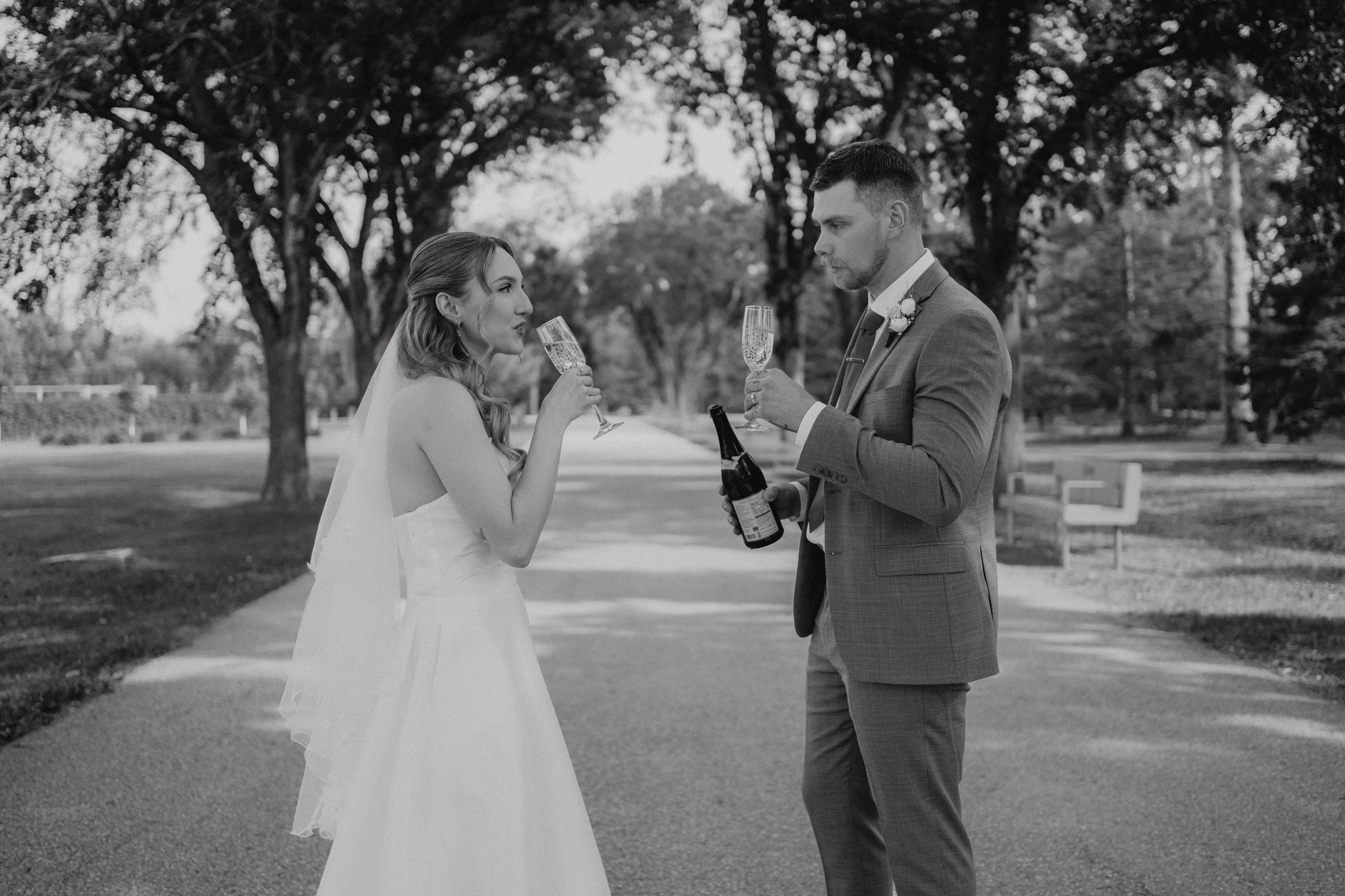Black and white photo of Liam and Tanya toasting champagne on the tree-lined path at Assiniboine Park — Winnipeg wedding photography by Ngo Photography