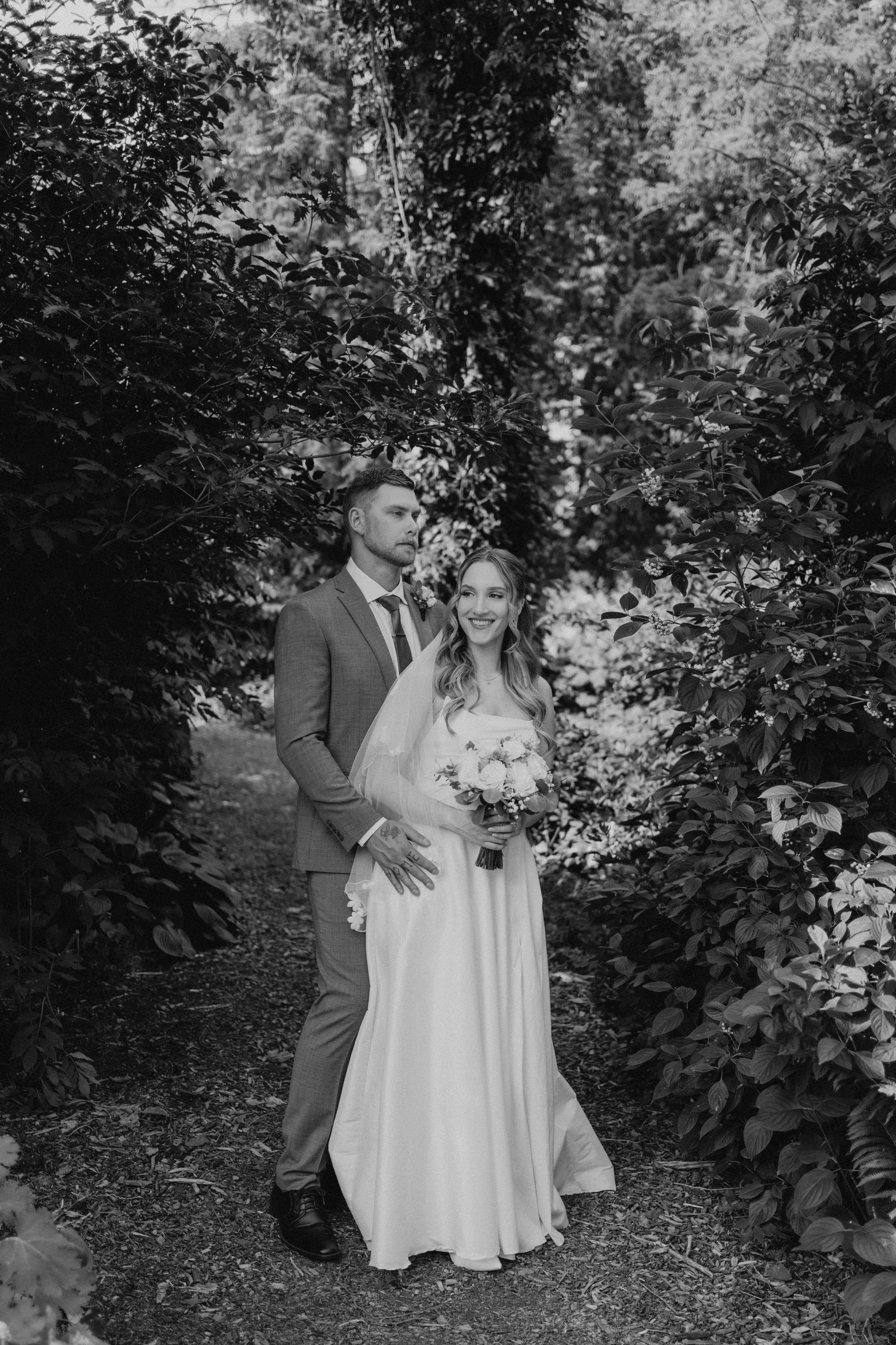 Black and white photo of Liam and Tanya walking hand in hand down a tree-lined garden path at Assiniboine Park — Ngo Photography