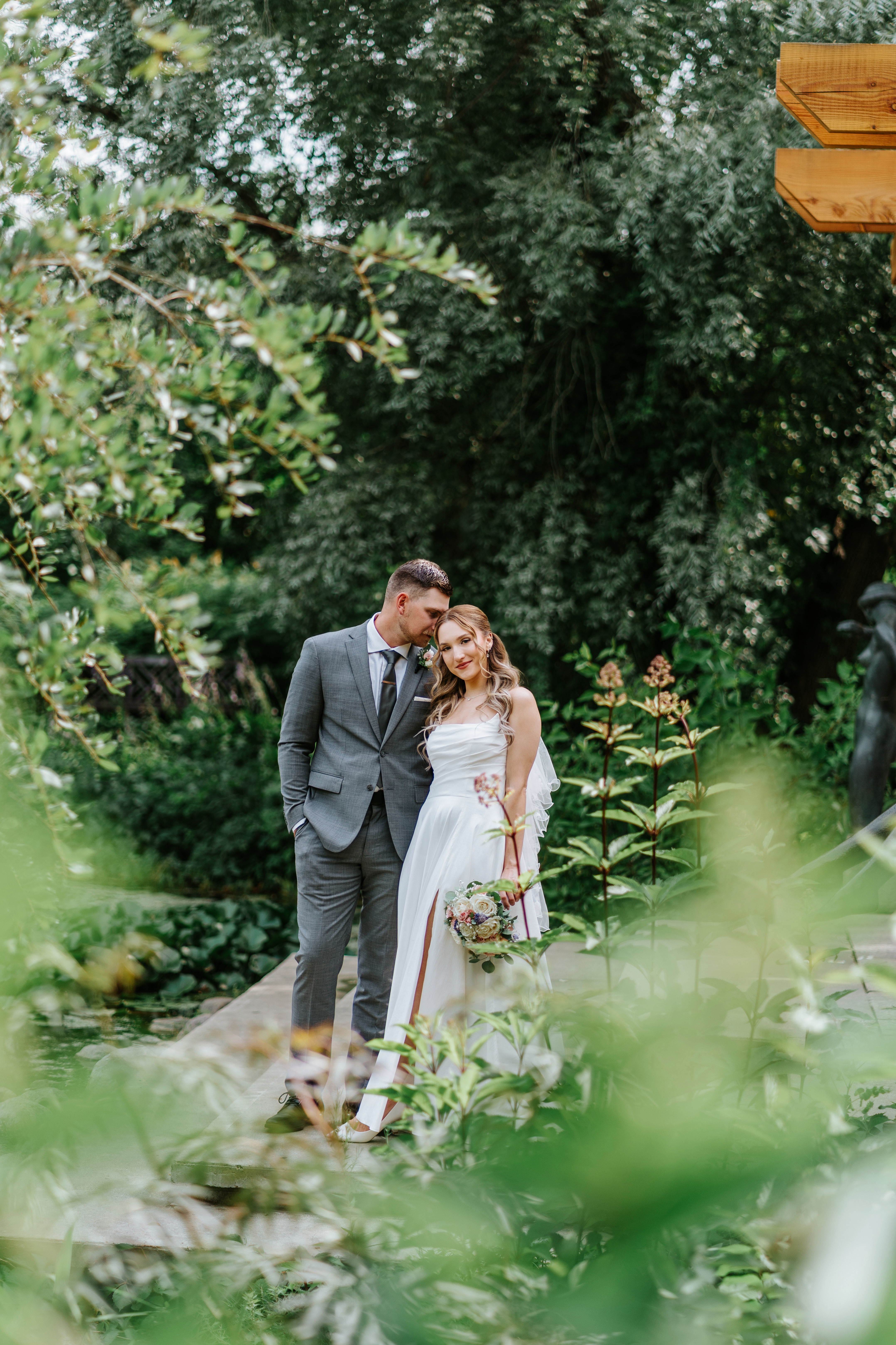 Liam and Tanya framed through lush garden foliage at Assiniboine Park — wedding portrait by Chris Ngo Winnipeg