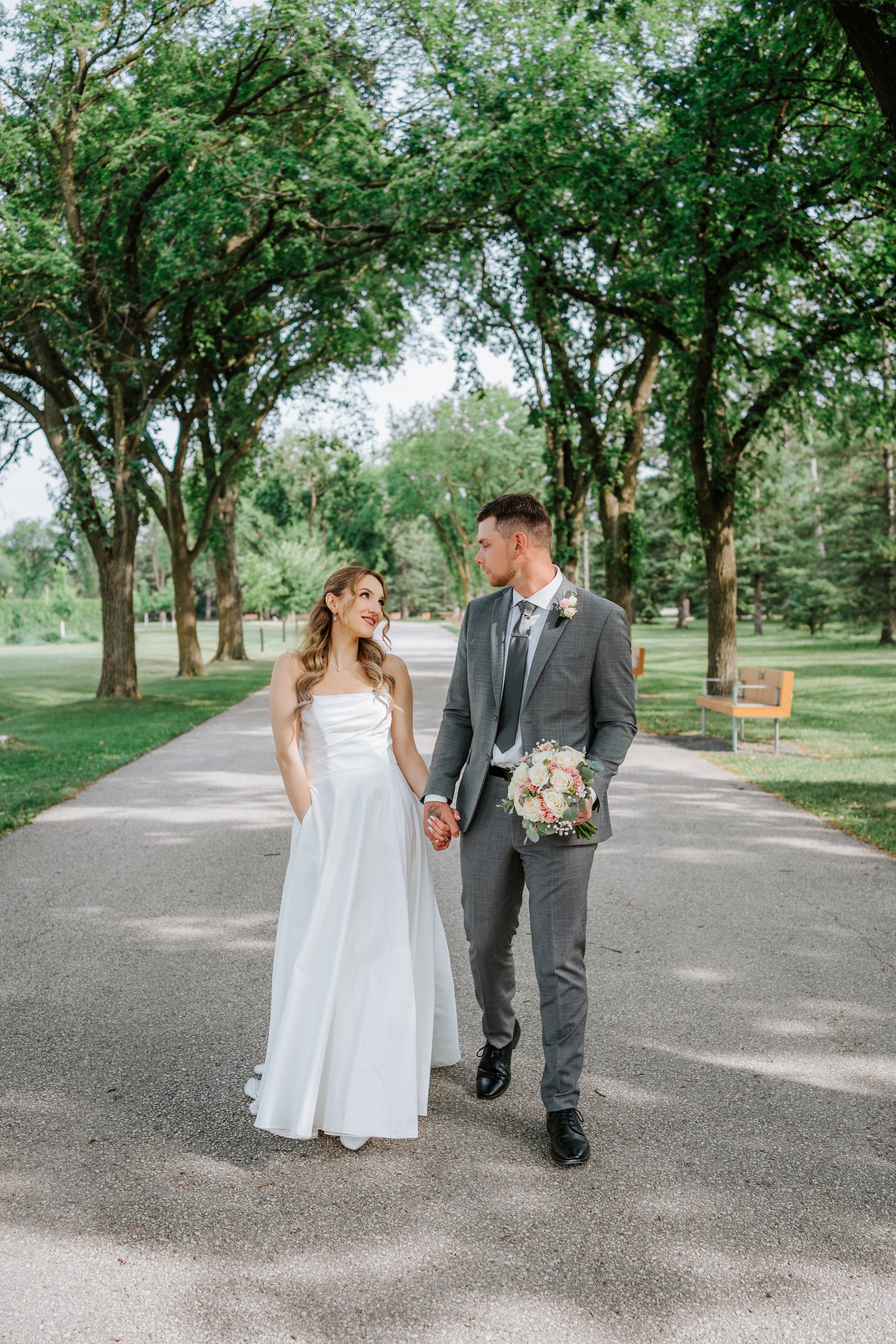 Liam and Tanya walking hand in hand and looking at each other on the Assiniboine Park avenue — summer wedding photography by Ngo Photography